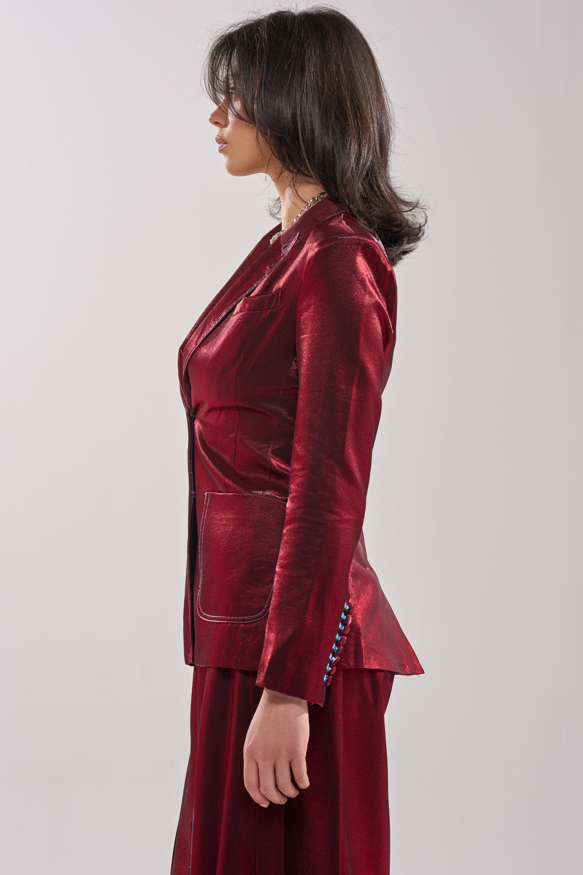 A woman with shoulder-length dark hair stands in profile, exuding power in a shiny red MISS YOU MOST BLAZER and matching pants against a plain light gray background. 2