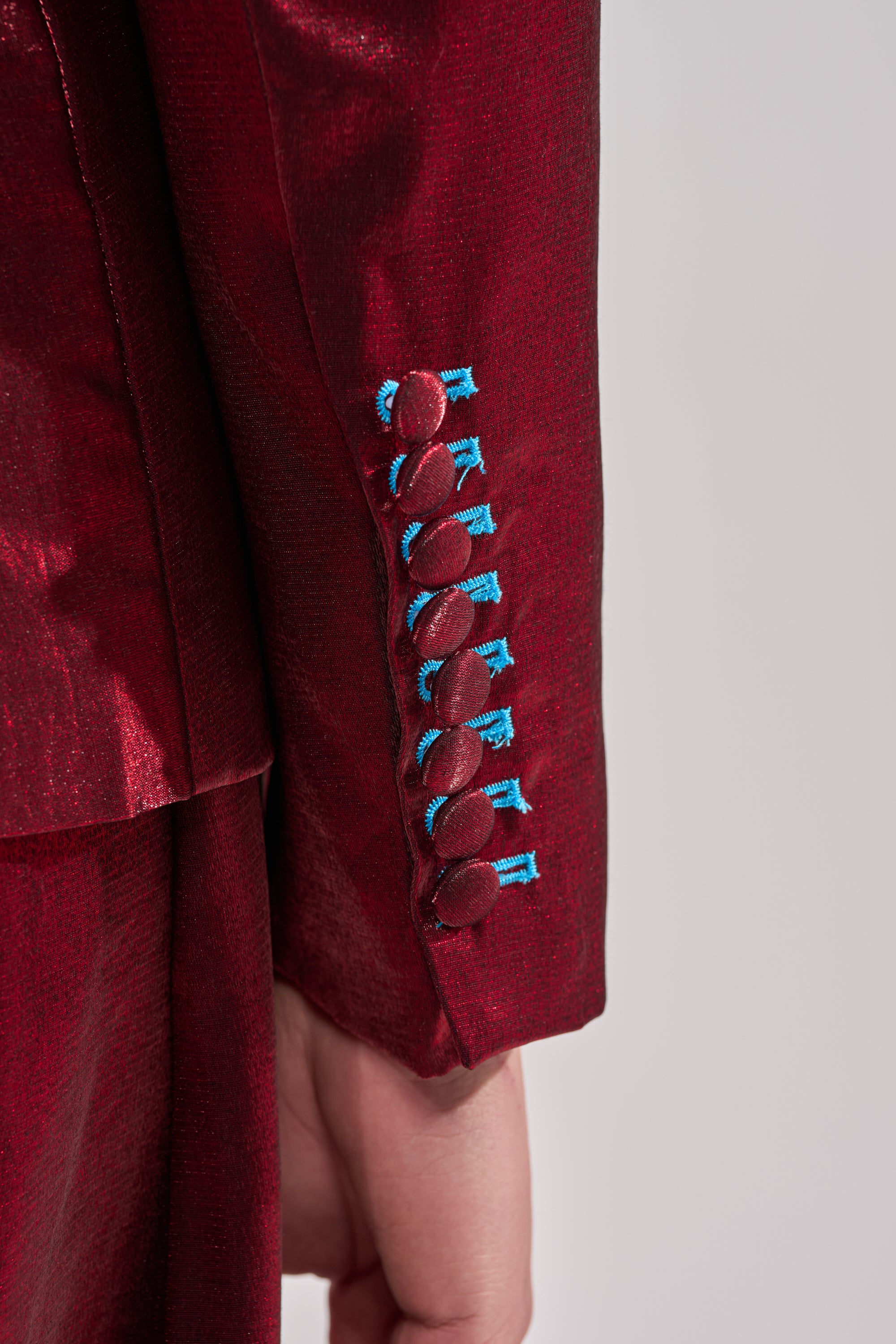 Close-up of a person's arm in the MISS YOU MOST BLAZER, featuring a shiny satin finish and maroon cuff buttons stitched with bright blue thread, set against a plain light background. 5