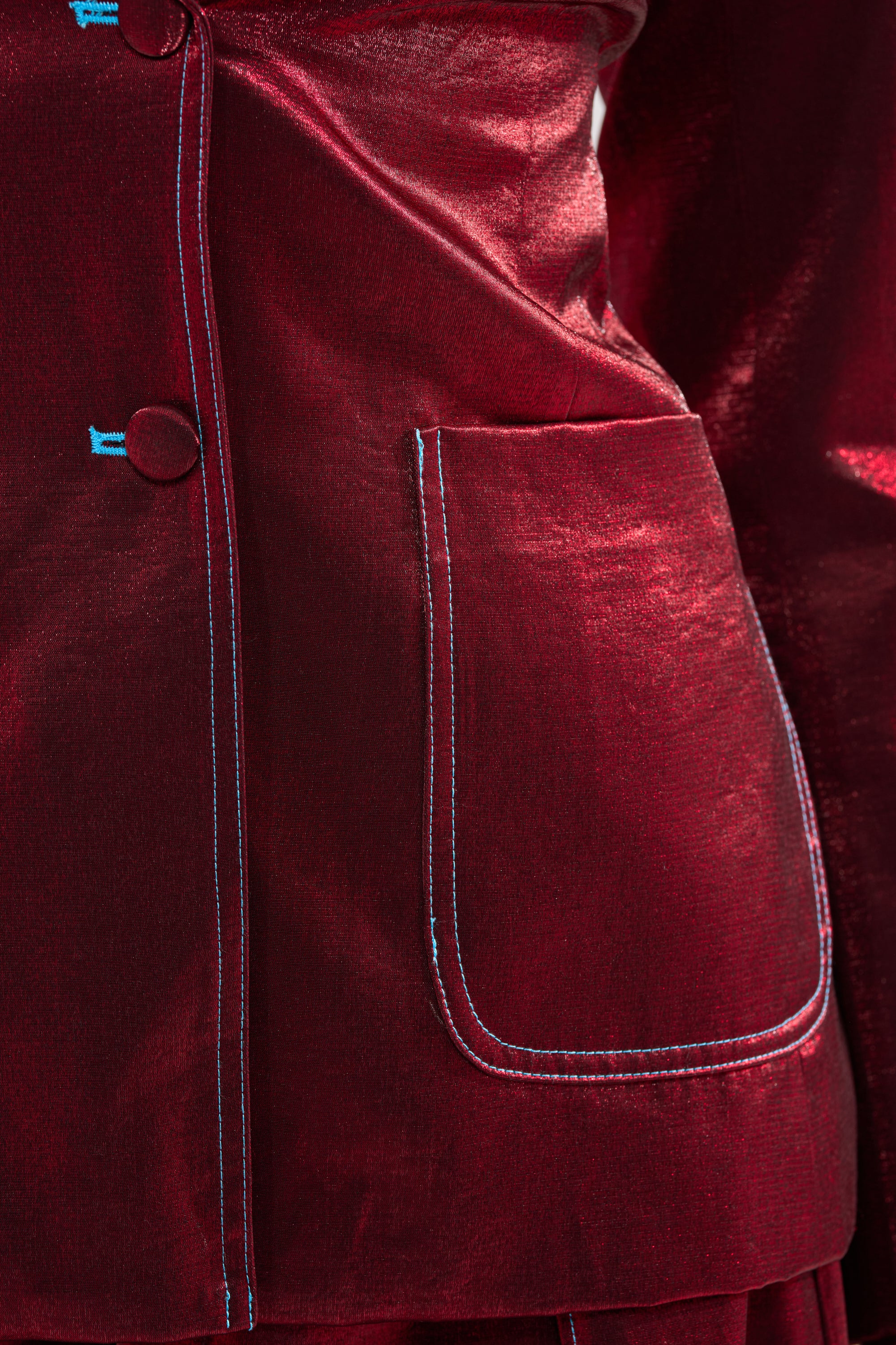 Close-up of the MISS YOU MOST BLAZER—a shiny red velvet blazer with a satin finish, light blue stitching, a large front pocket, and two buttons along the seam. 6