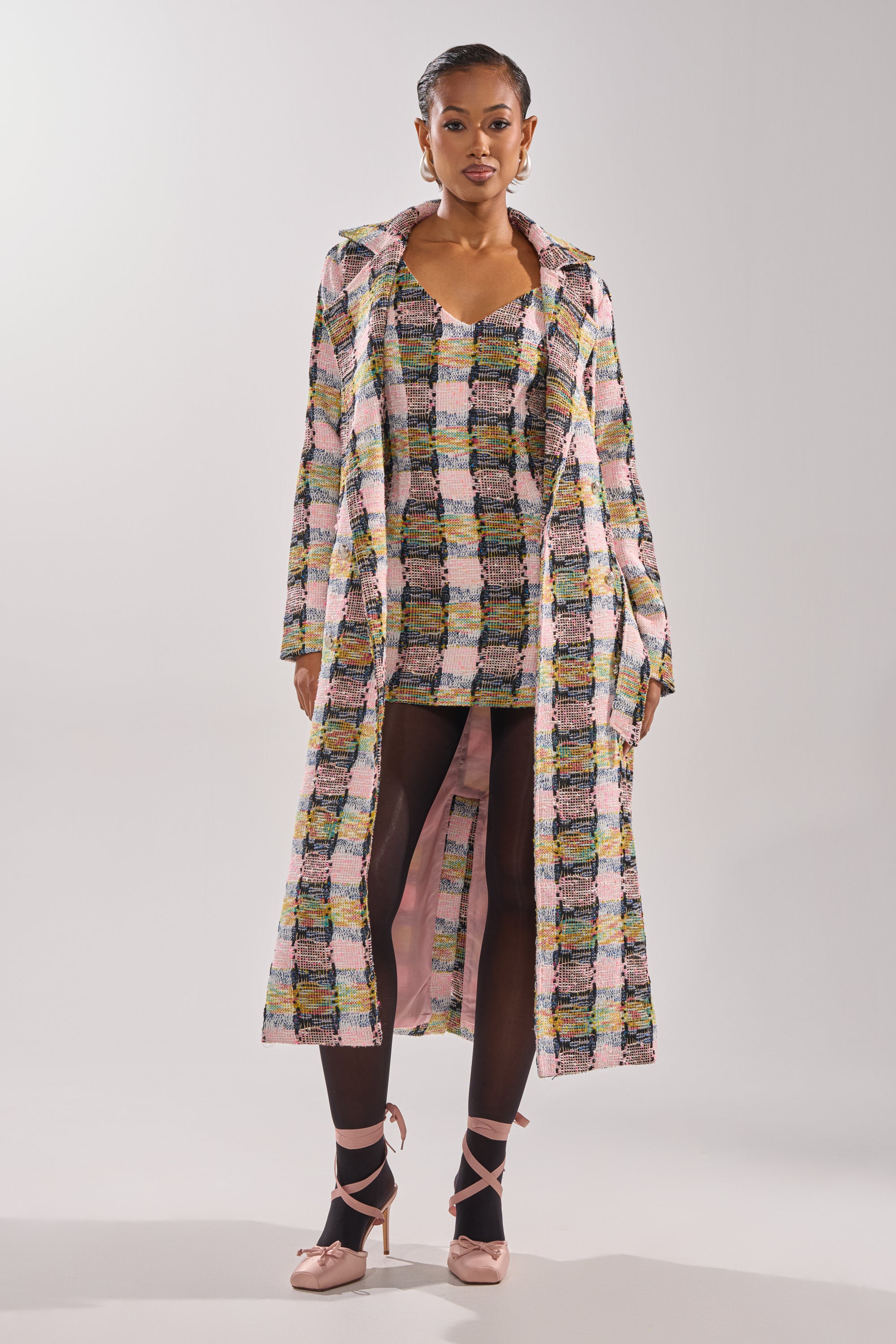 A woman faces forward in the MAIN SQUEEZE TWEED TRENCH, a long pastel check coat over a matching dress. She wears dark tights and pink ballet flats with her hair styled back, set against a plain, light background. 1