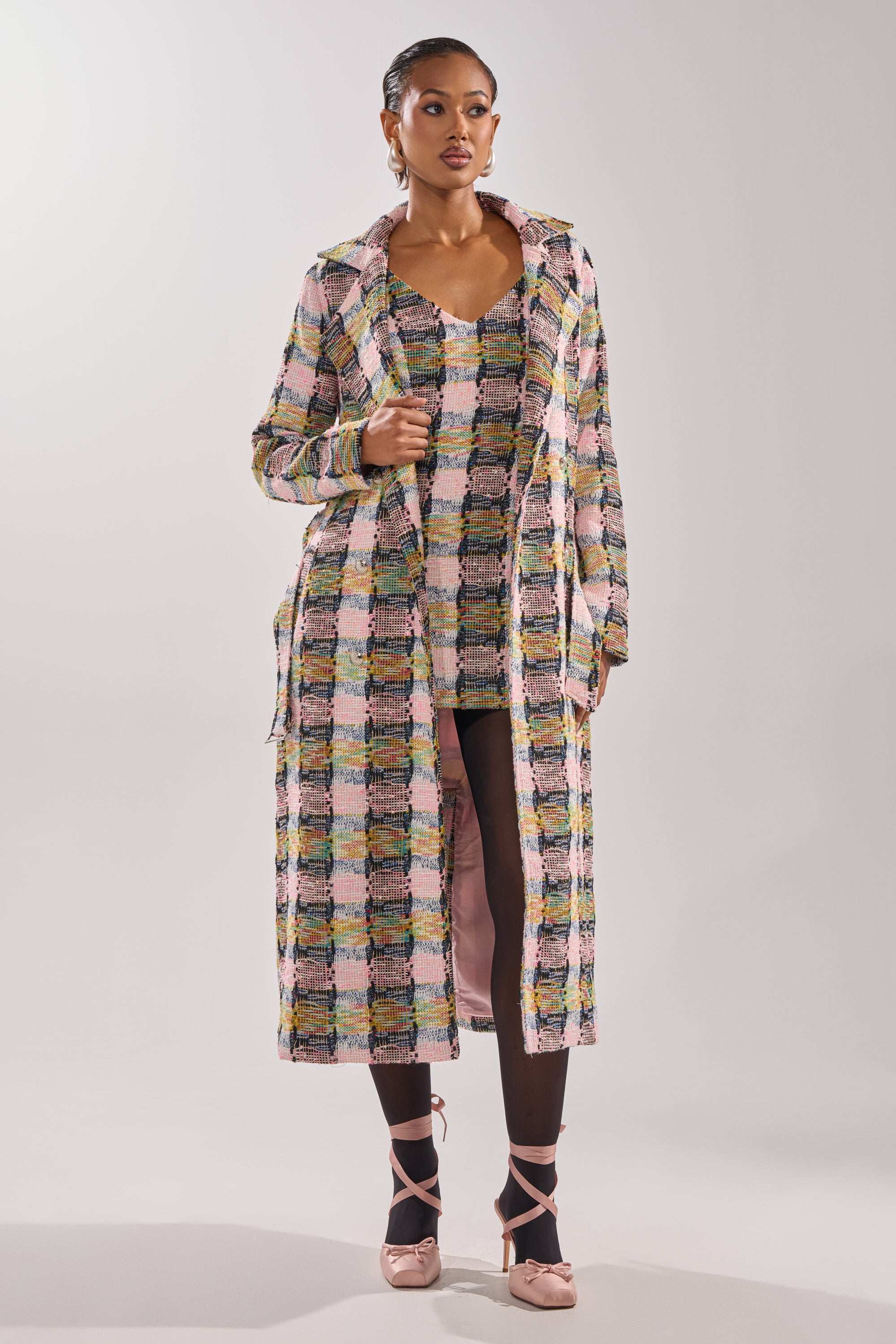 A woman poses confidently against a plain backdrop wearing the MAIN SQUEEZE TWEED TRENCH over a matching dress, styled with sheer black tights and blush pink ballet flats. 2