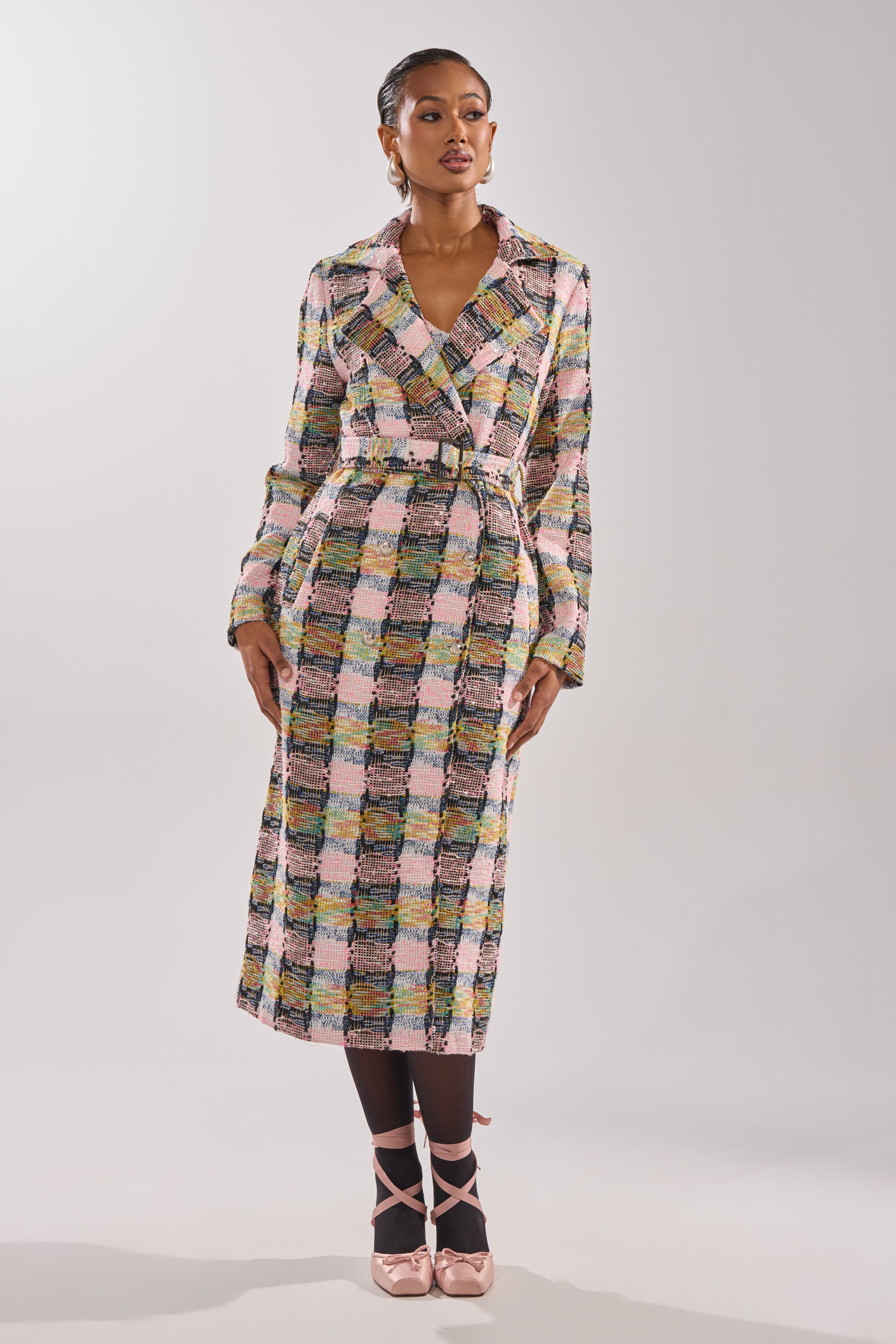 A woman poses against a plain light background wearing the AKIRA Label MAIN SQUEEZE TWEED TRENCH, a pastel check belted coat with a wide collar, styled with black tights and pale pink ballet flats. 3