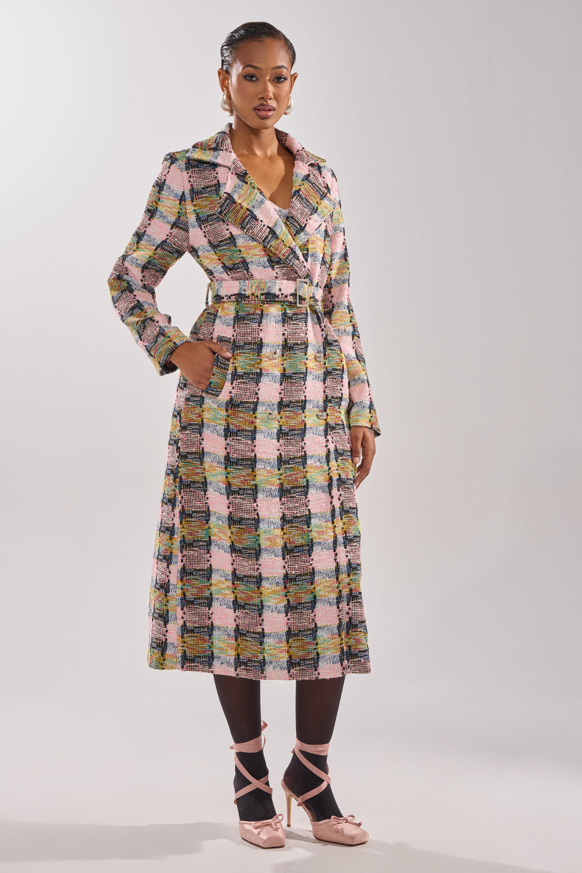 A woman wears the MAIN SQUEEZE TWEED TRENCH, a pastel check AKIRA Label coat with notched collar and belt. She pairs it with black tights and pink ballet flats, standing against a plain light gray background. 4