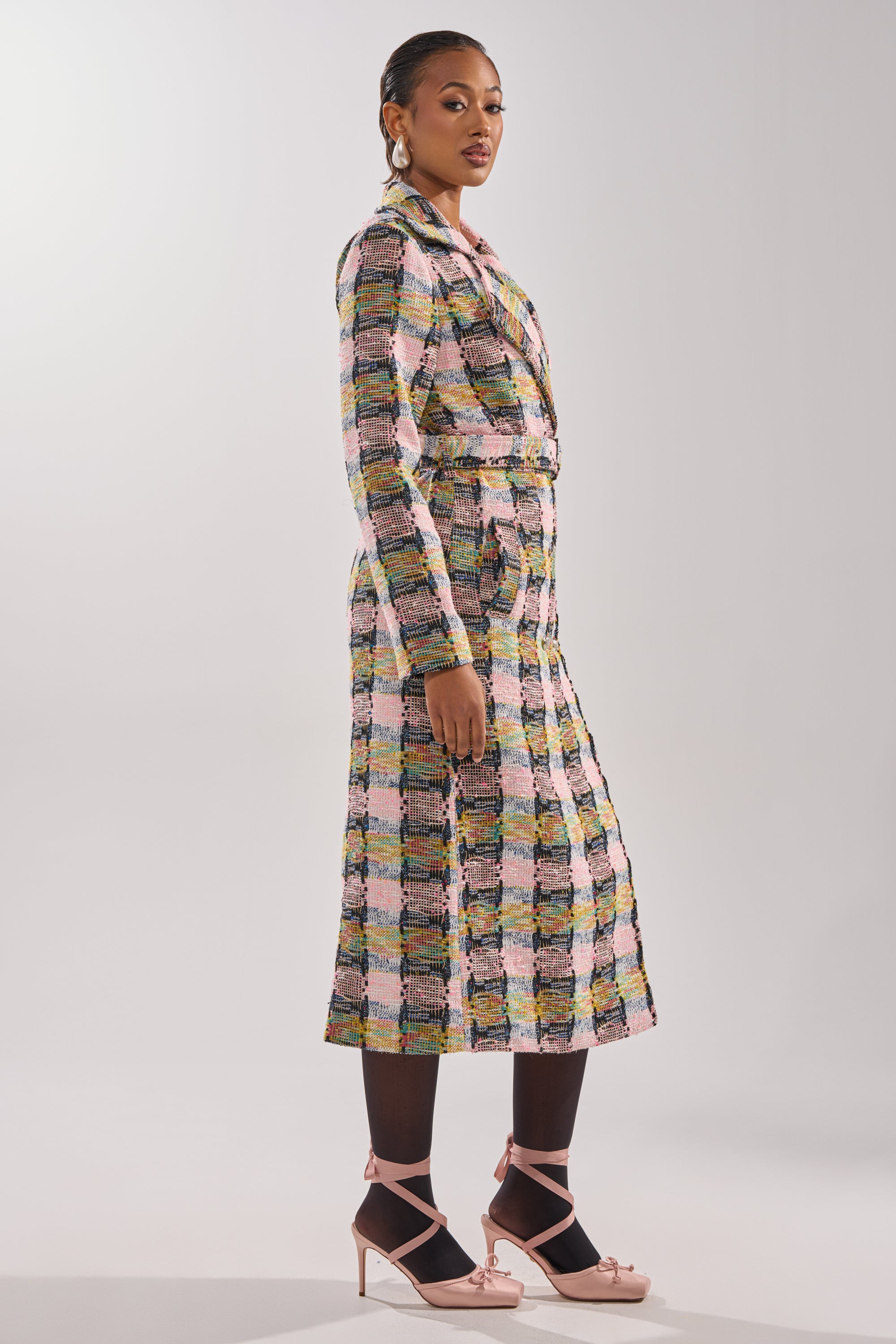 A woman in profile wears the MAIN SQUEEZE TWEED TRENCH with black tights, pink ankle-strap heels, and gold hoop earrings. Her hair is tied back, and she stands against a simple light backdrop. 5
