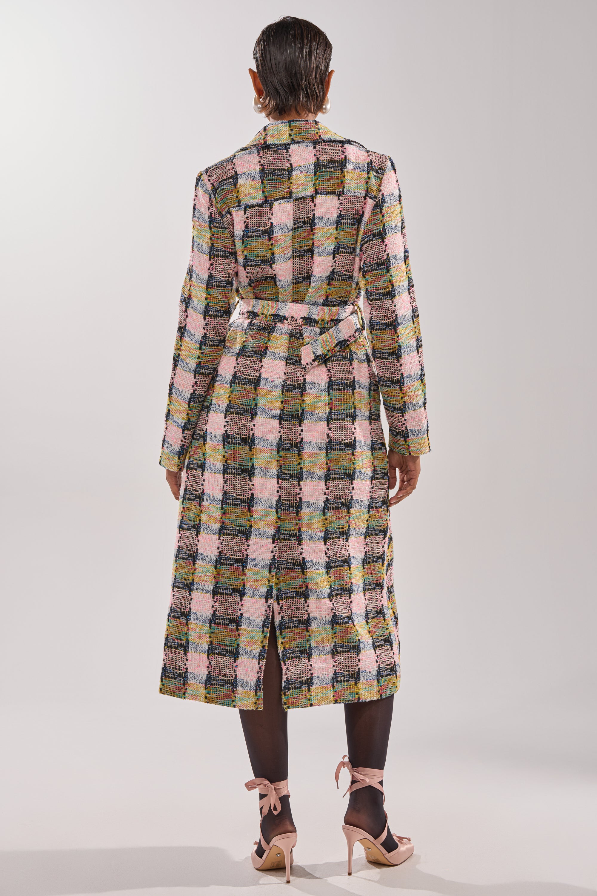 A person with short dark hair faces away, wearing the MAIN SQUEEZE TWEED TRENCH. She pairs it with sheer black stockings and beige high-heeled sandals with ankle bows for a chic look. 6