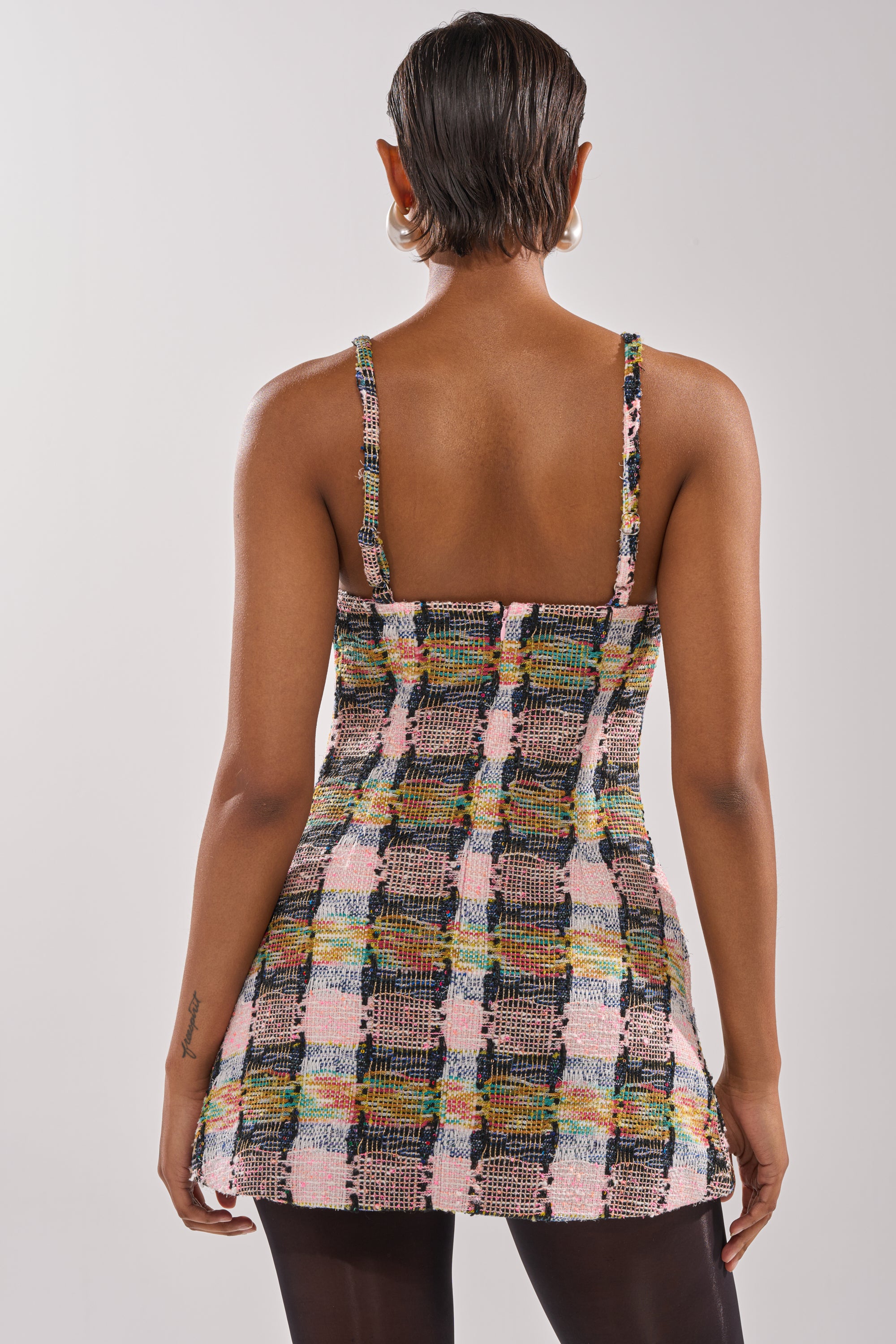 A person with short hair and hoop earrings wears the MAIN SQUEEZE TWEED MINI DRESS, a sleeveless multicolor plaid mini dress, seen from the back against a plain light background. 3