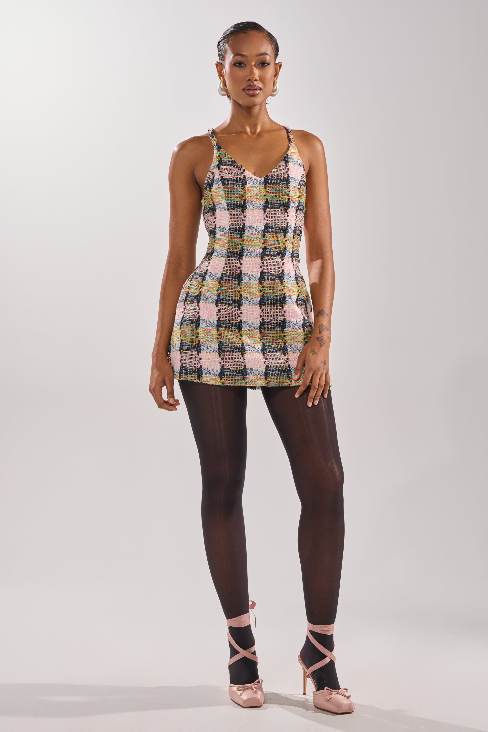 A woman stands against a plain background, confidently looking at the camera in the MAIN SQUEEZE TWEED MINI DRESS, styled with black sheer tights and pink high-heeled ankle strap shoes. 4