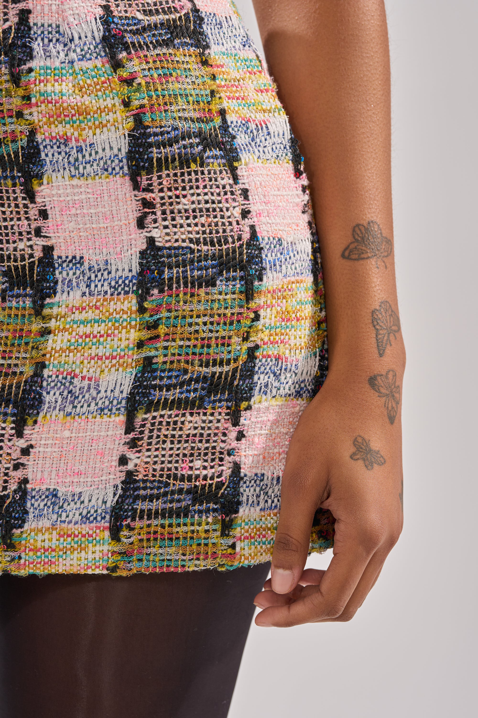 A close-up of a person's arm with butterfly tattoos beside the MAIN SQUEEZE TWEED MINI DRESS, featuring a colorful pink, blue, yellow, and black plaid pattern against a plain, neutral background. 7