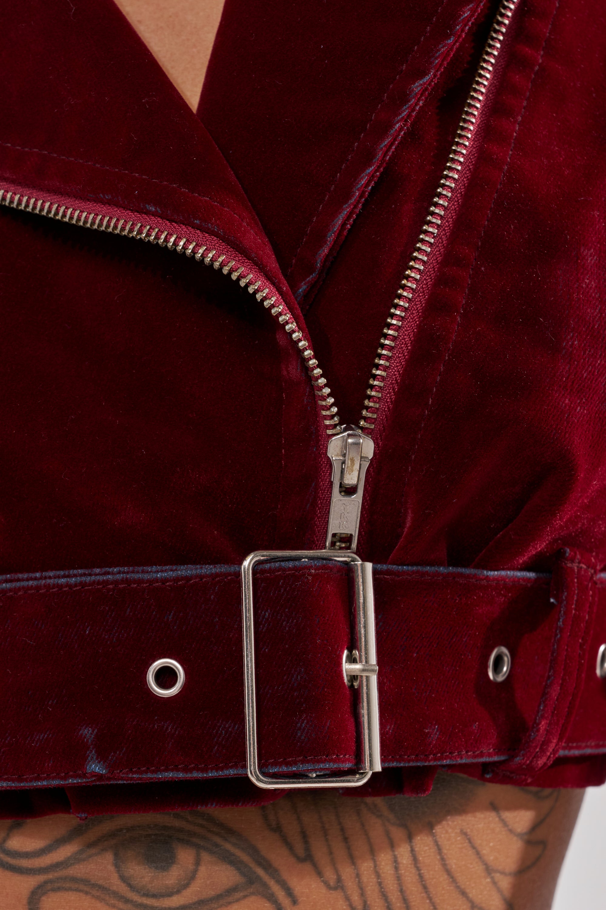 Close-up of the LULU VELVET DENIM MOTO jacket in red, featuring a silver zipper and belt buckle; a tattooed arm with a stylized eye design appears at the bottom.