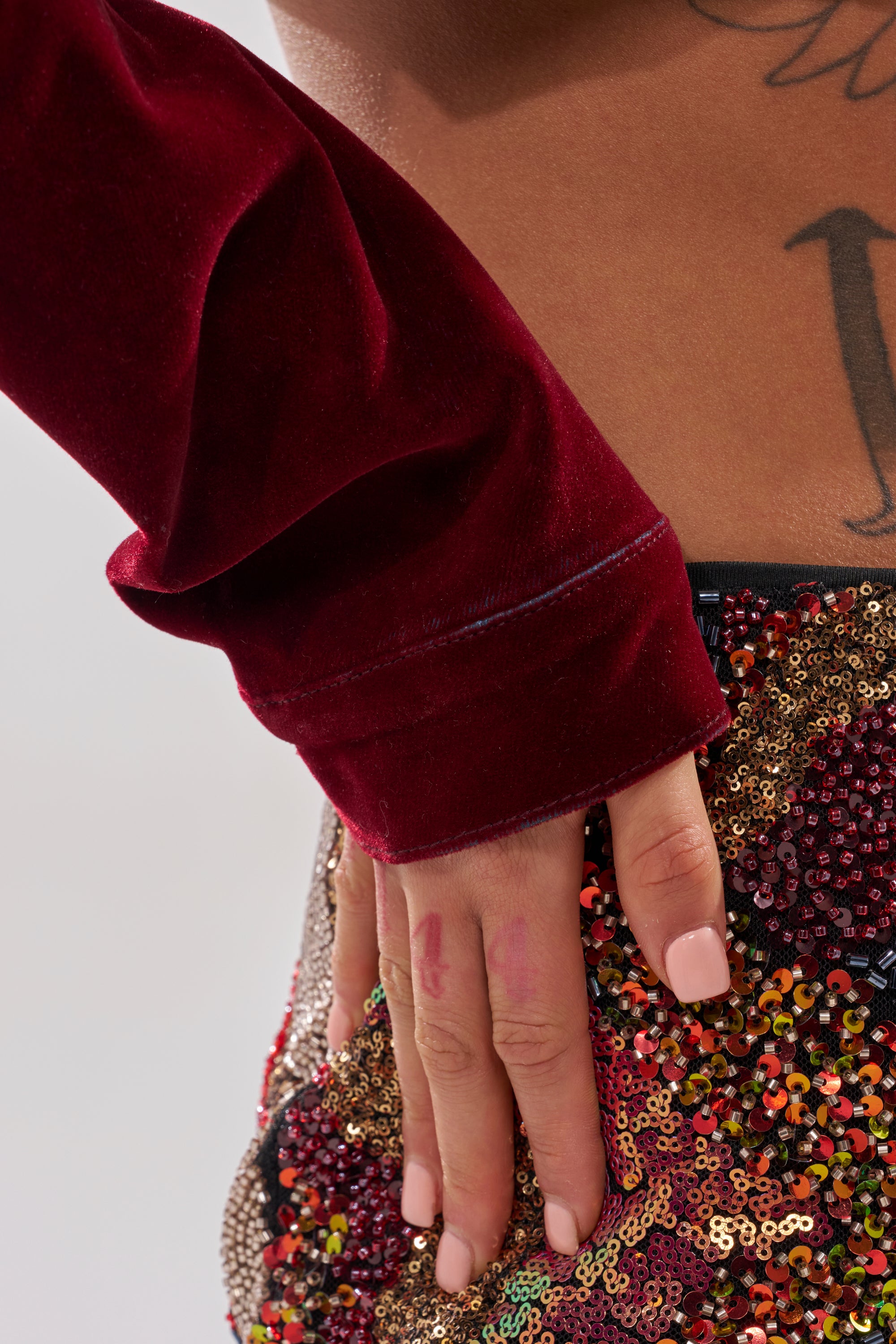 A close-up of a hand on a sequined, multicolored skirt. The person wears the LULU VELVET DENIM MOTO jacket in red wine with long sleeves, and a tattoo is visible on their bare back above the waistband.