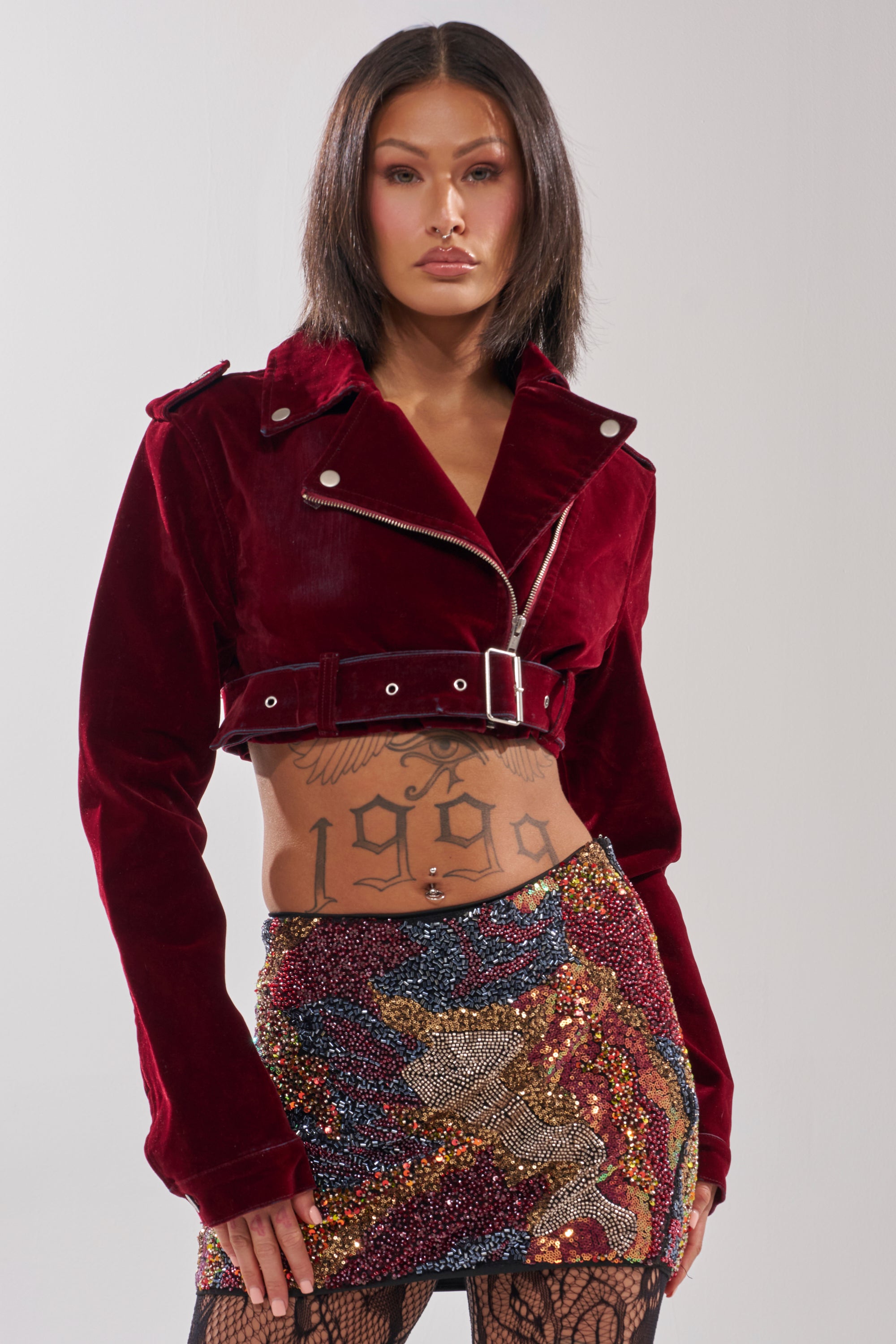 A woman with straight, dark hair wears the LULU VELVET DENIM MOTO jacket, paired with a multicolored sequined mini skirt. She has torso tattoos and neutral-toned makeup.