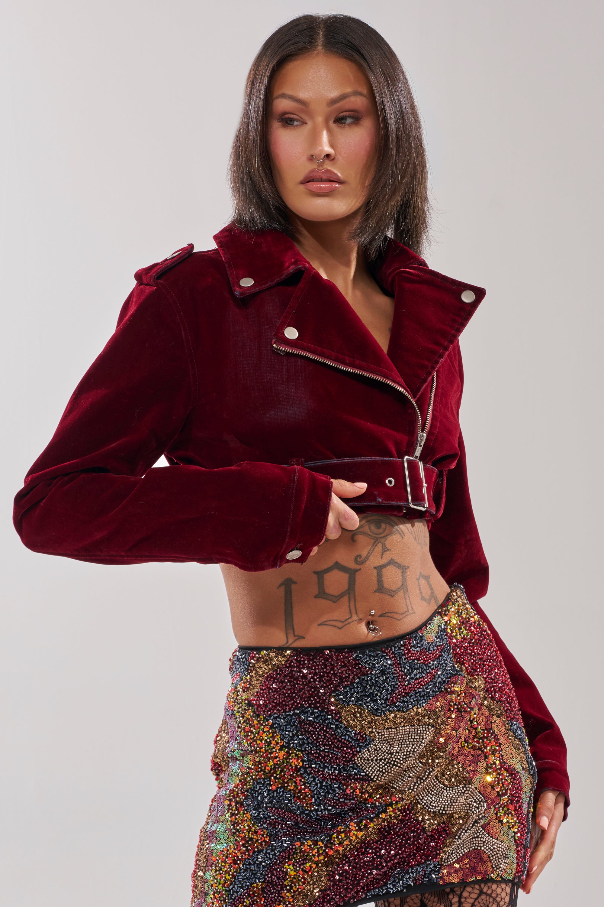 A person with straight, shoulder-length hair wears the LULU VELVET DENIM MOTO in burgundy with silver buttons and a colorful sequined mini skirt. A "1999" tattoo is visible on their torso as they look off to the side.