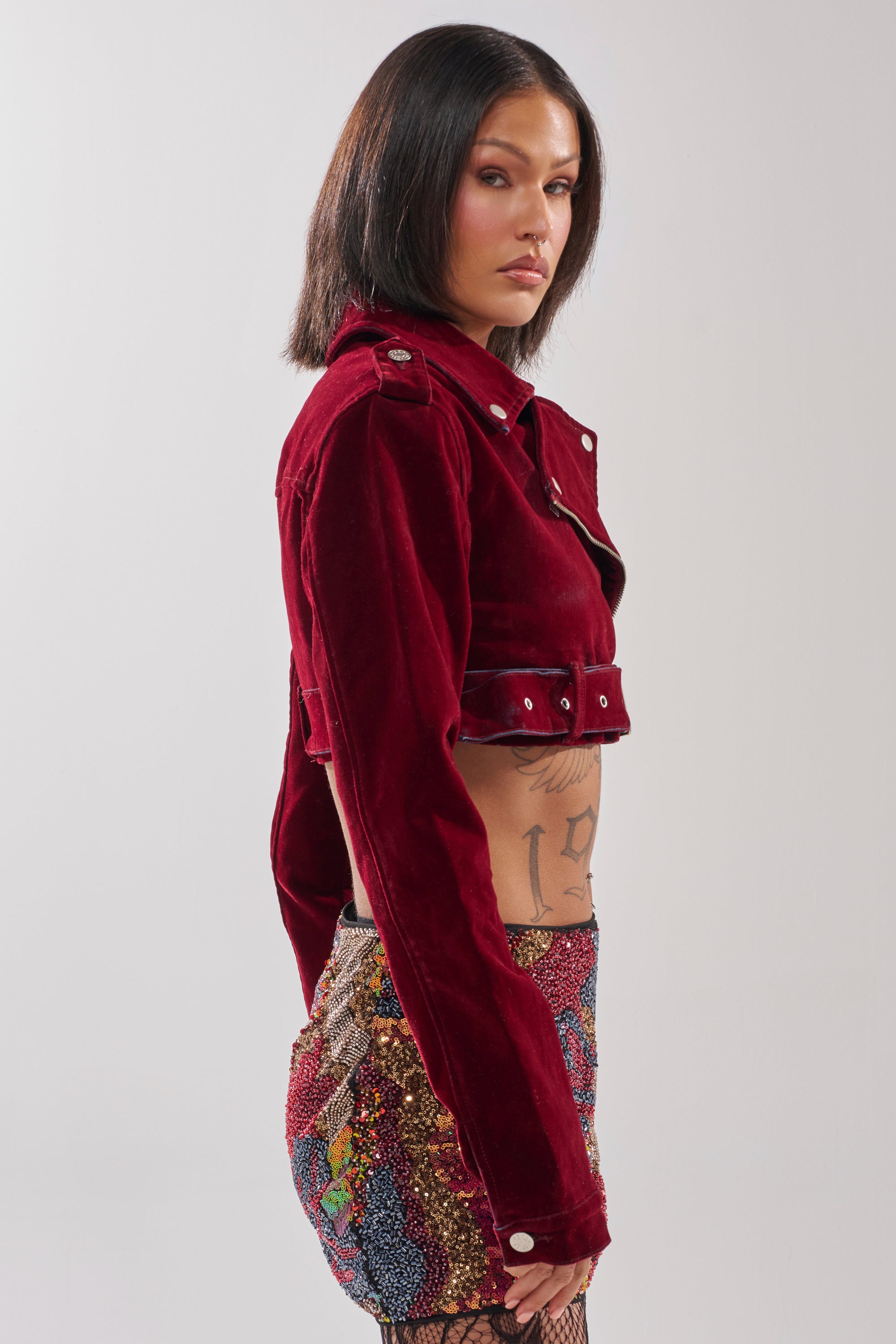 A woman with straight dark hair and visible ribcage tattoos wears the LULU VELVET DENIM MOTO jacket in red velvet, paired with a colorful sequin mini skirt. She stands sideways against a plain light background.