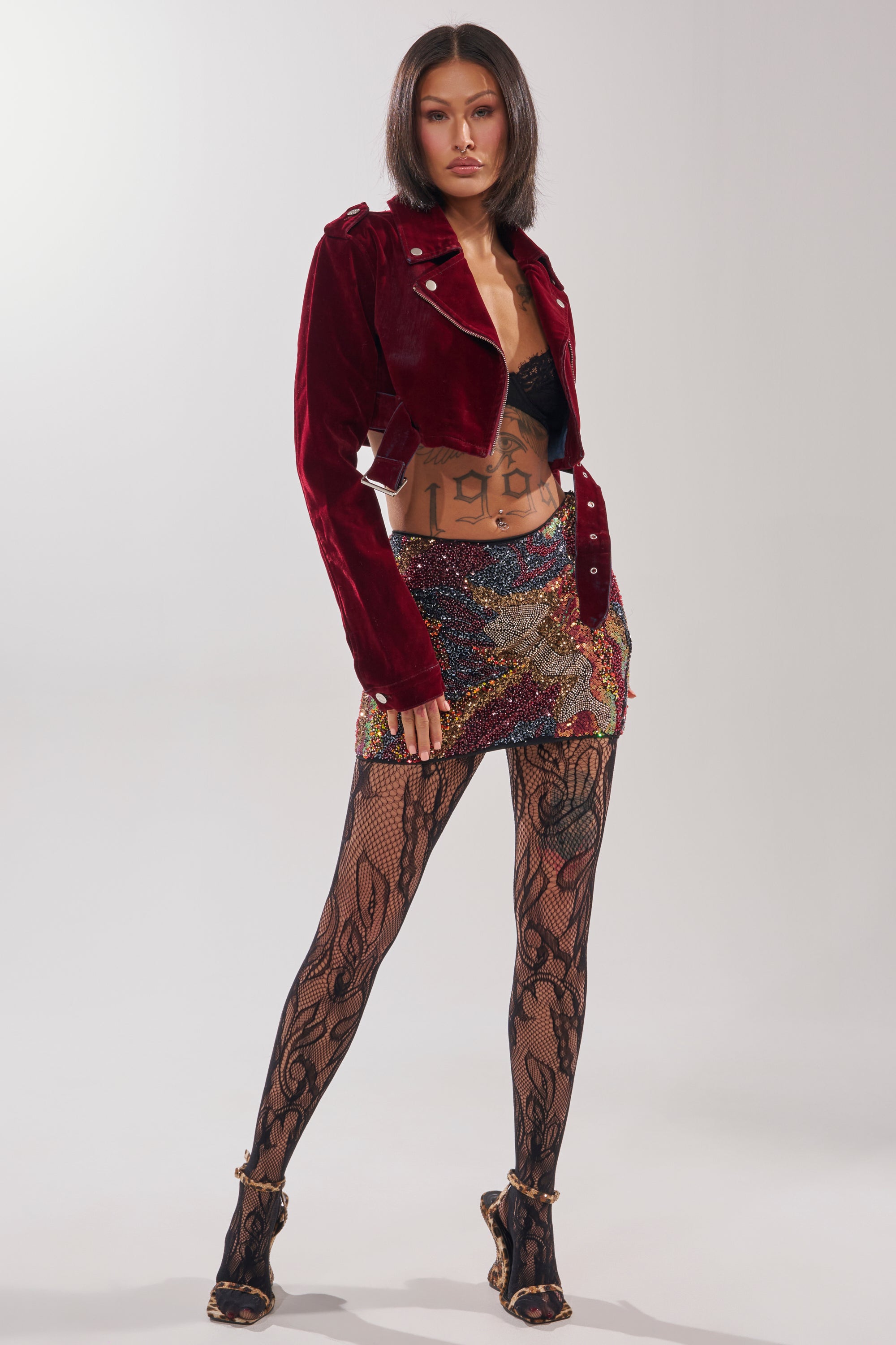 A woman with a bob haircut stands confidently against a plain light background, wearing the LULU VELVET DENIM MOTO jacket, a black lace bra, multicolored sequin mini skirt, patterned lace tights, and high heels.
