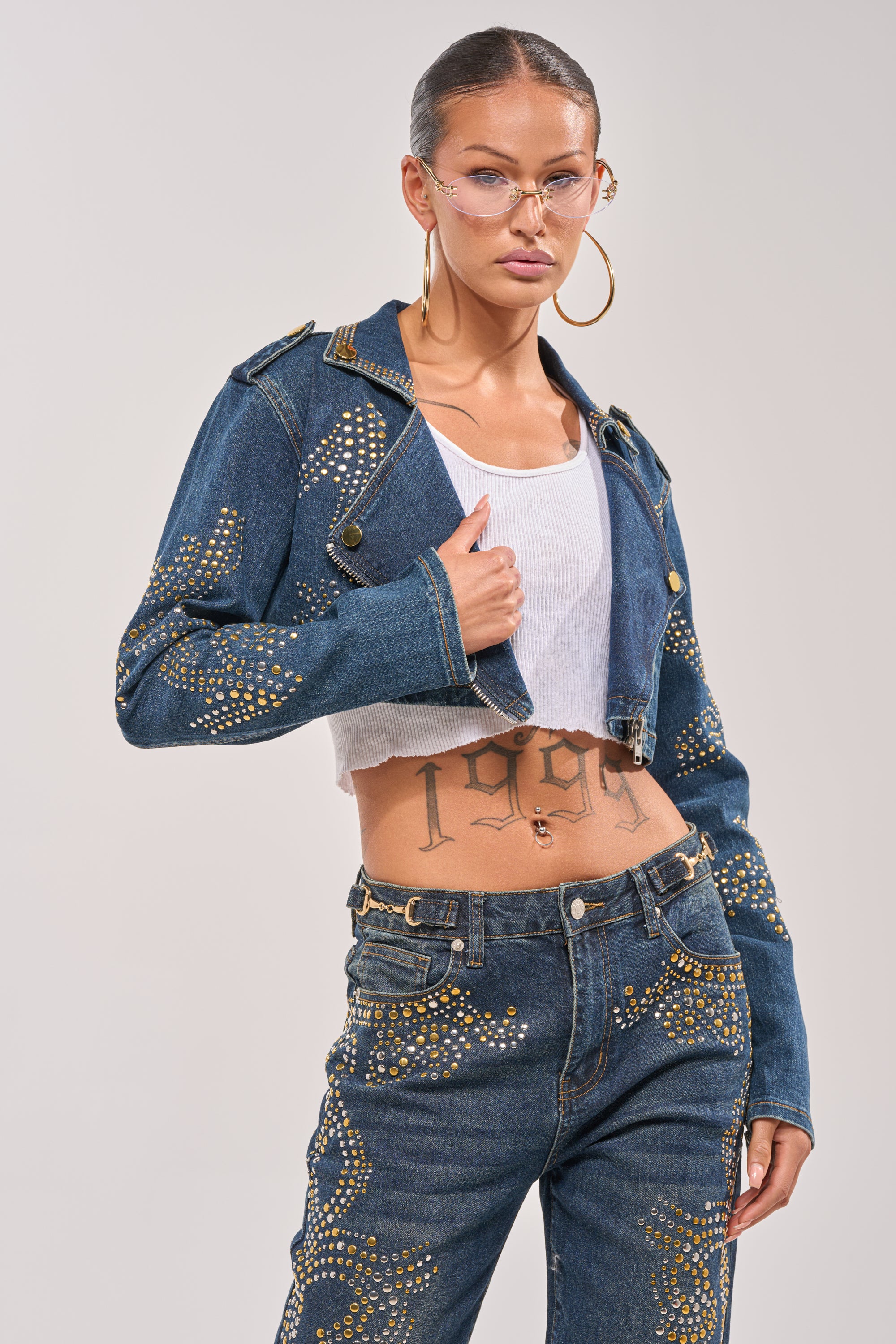Wearing the OFF TO THE RODEO CROPPED MOTO jacket, matching denim pants, hoop earrings, and gold-rimmed glasses, a woman confidently poses in a white crop top, flashing her "1999" tattoo against a plain background. 0