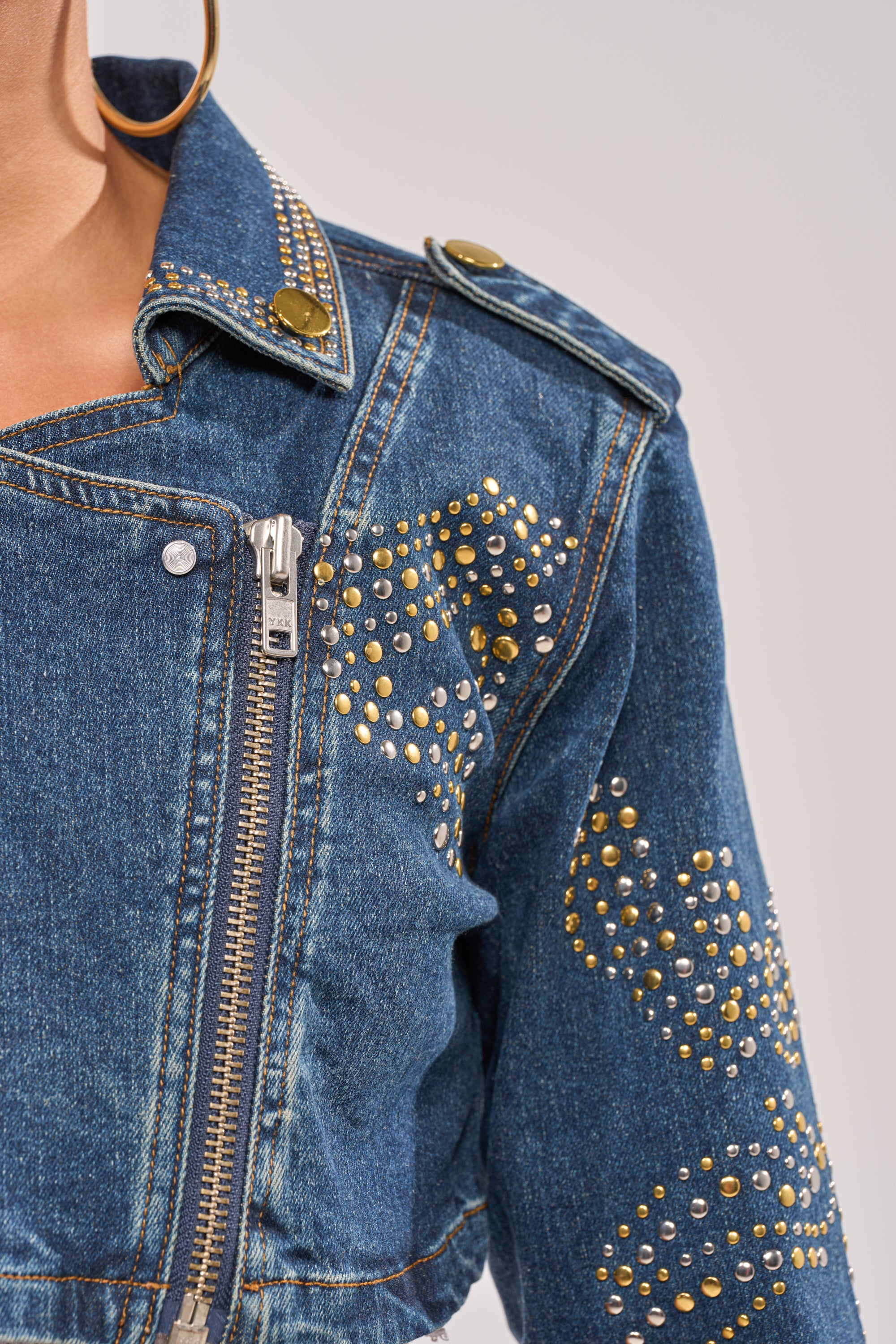Close-up of the OFF TO THE RODEO CROPPED MOTO denim jacket with gold and silver studs in patterns, zipper closure, metal collar button, epaulet detail, and a visible gold hoop earring. 9