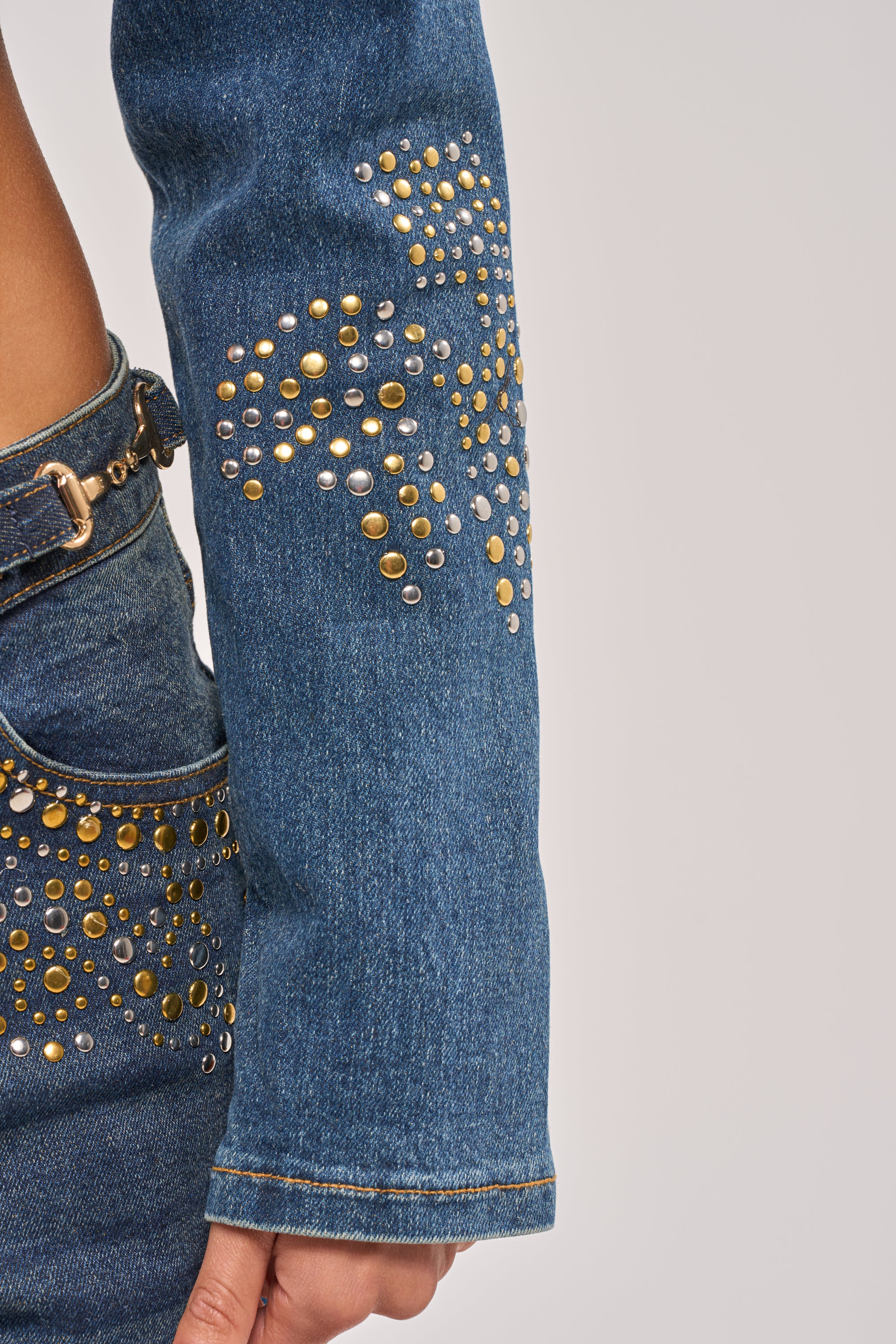 A close-up of a person's arm in the OFF TO THE RODEO CROPPED MOTO, a blue denim jacket with gold and silver studs in striking patterns. Matching denim pants feature similar studs for the ultimate festival-ready look. 10