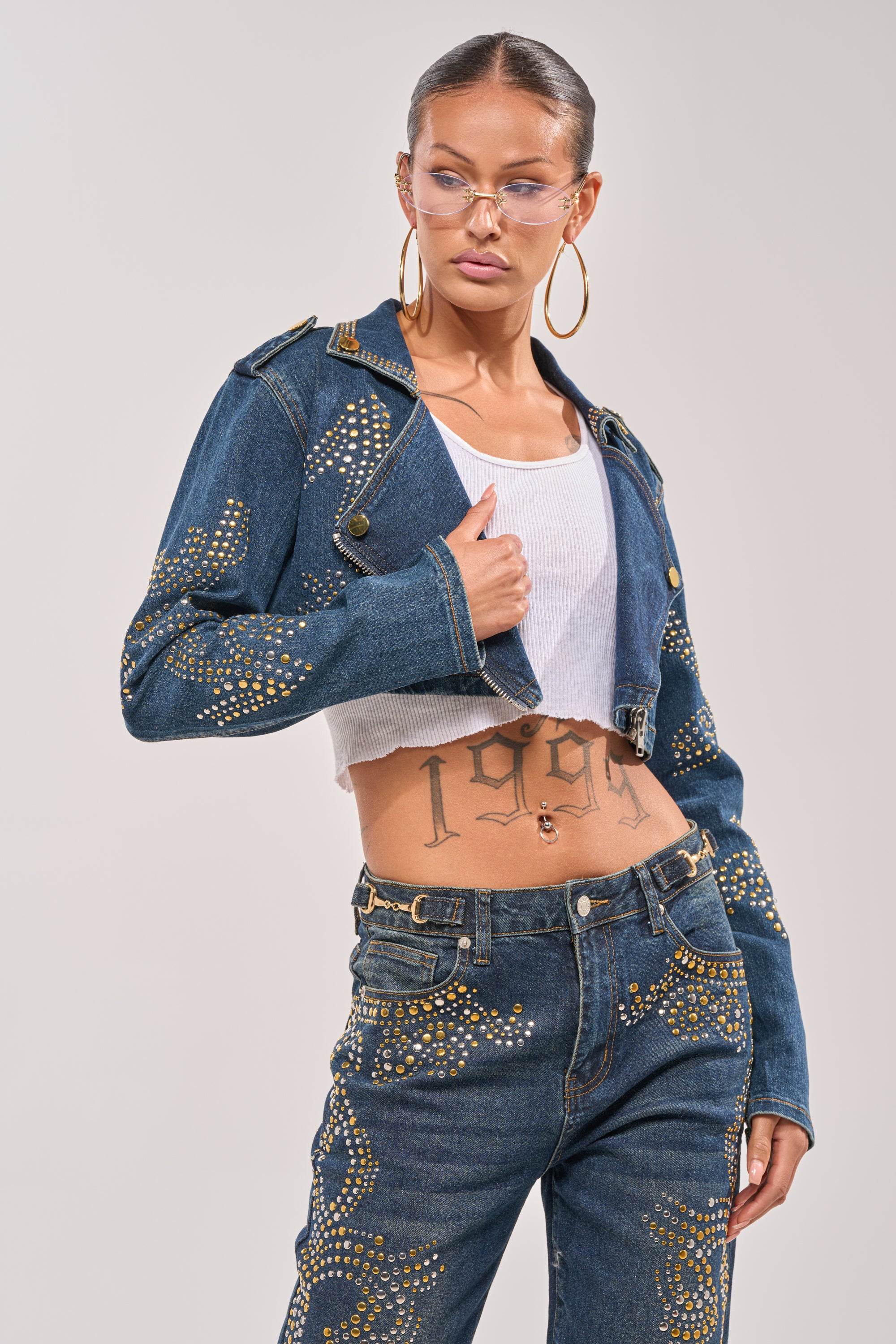 A person wears the OFF TO THE RODEO CROPPED MOTO studded denim jacket with jeans featuring gold studs, a white tank top, large hoop earrings, clear glasses, slicked-back hair, and a visible "1994" stomach tattoo. 1