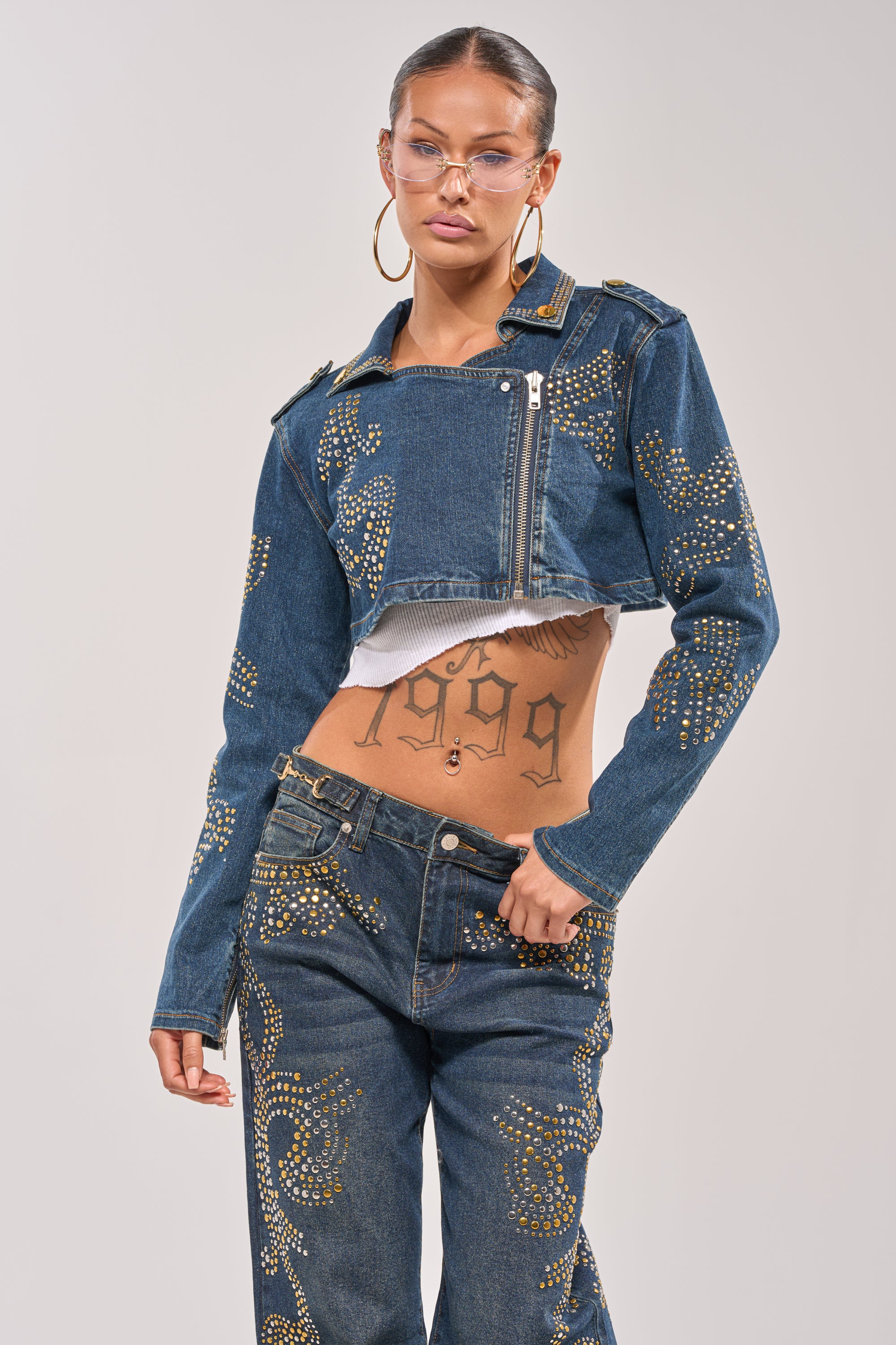 Wearing the OFF TO THE RODEO CROPPED MOTO jacket and matching jeans with gold accents, clear glasses, large hoop earrings, and a “1999” stomach tattoo. 2