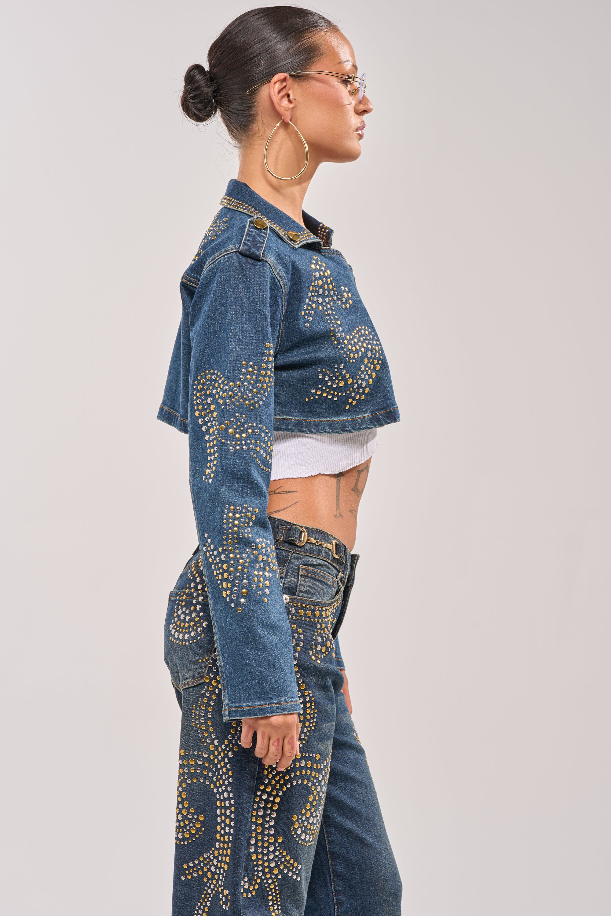 A woman in profile wears the OFF TO THE RODEO CROPPED MOTO jacket with gold-studded jeans, a white crop top, hoop earrings, sunglasses, and a sleek bun, showing a tattoo on her waist and torso. 3