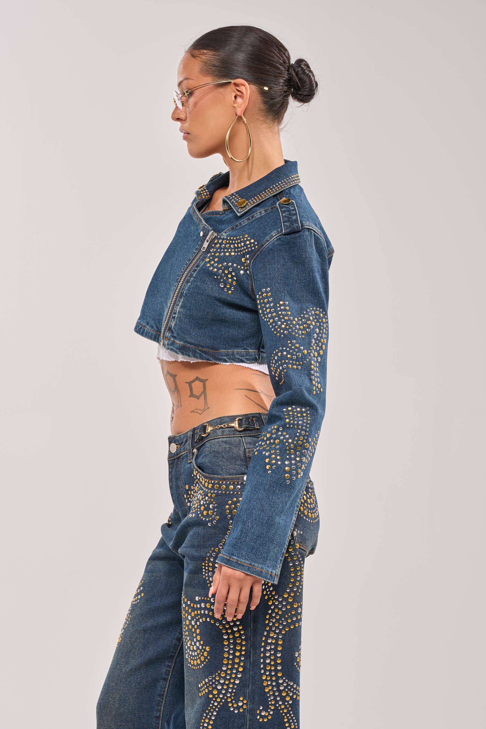 A woman in profile wears the OFF TO THE RODEO CROPPED MOTO jacket and matching jeans with gold accents, styled with a bun, hoop earrings, glasses, and a visible stomach tattoo. 4
