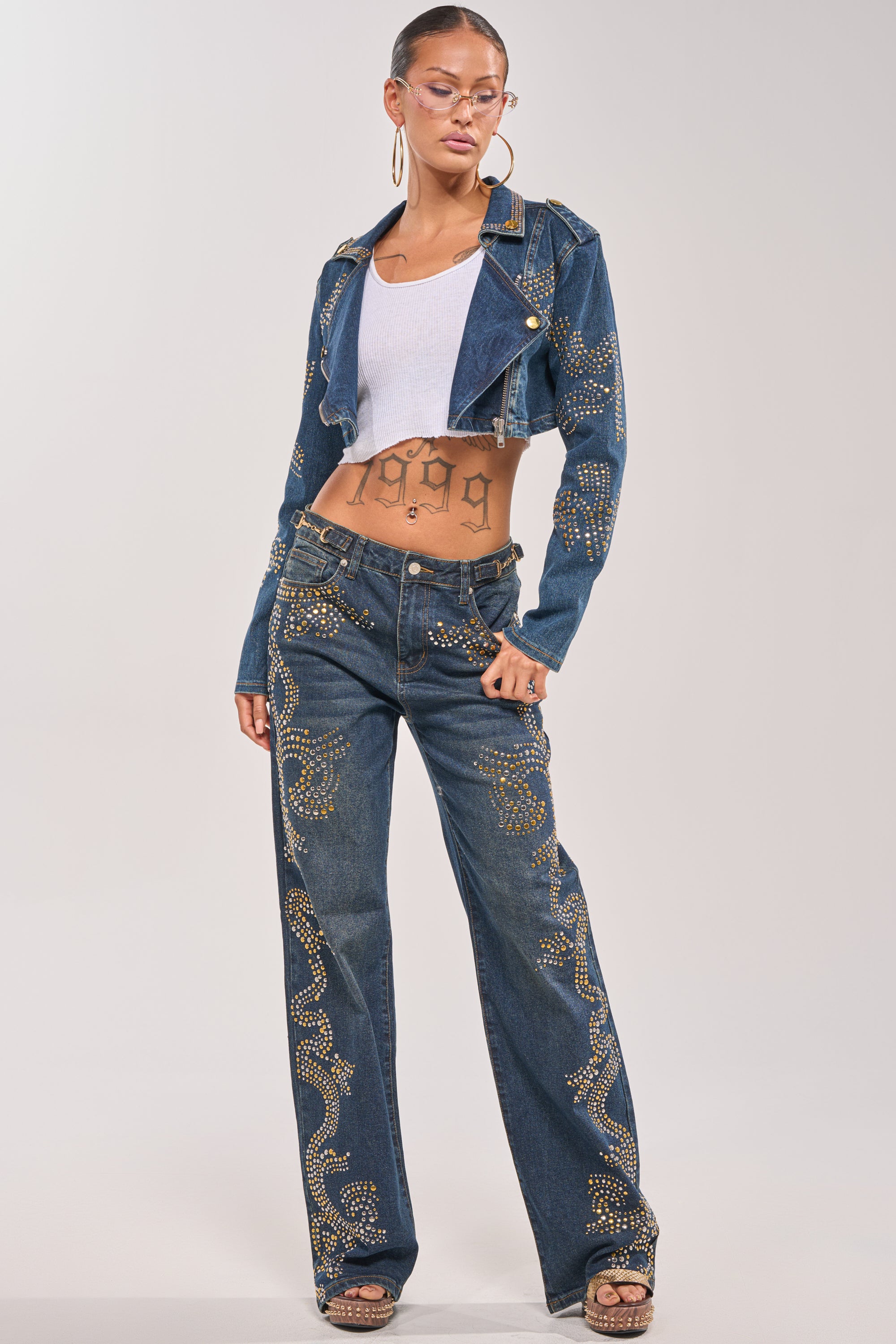 A woman stands confidently in the OFF TO THE RODEO CROPPED MOTO, a studded denim moto jacket with matching jeans featuring swirling gold studs. She pairs it with a white crop top, hoop earrings, glasses, and a "1999" tattoo on her stomach. 6