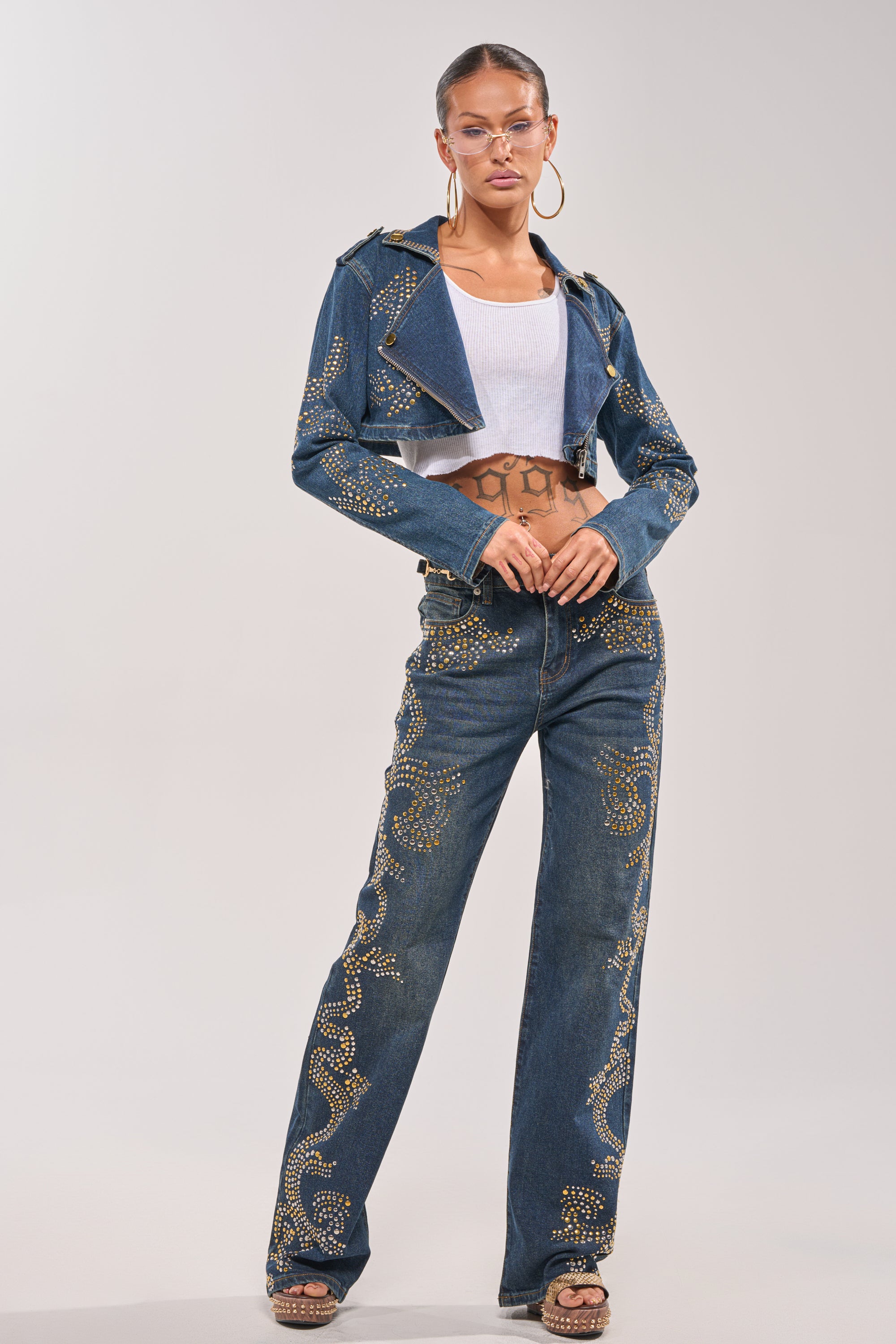 A woman poses in the OFF TO THE RODEO CROPPED MOTO blue studded denim jacket and matching high-waisted jeans with gold accents, styled with a white crop top, hoop earrings, clear glasses, and platform shoes against a plain background. 8