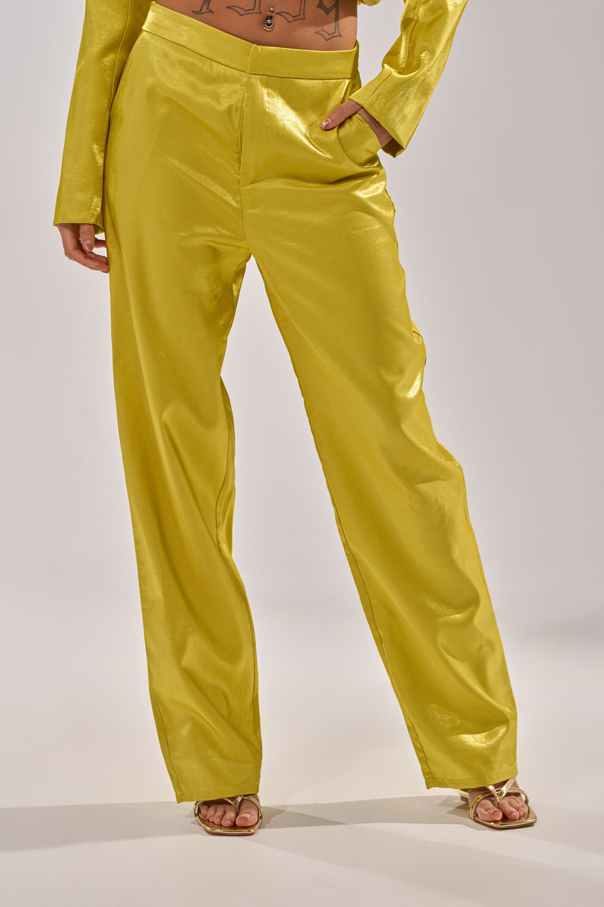 A person wears the CLEO TROUSER IN CHARTREUSE, featuring a shiny finish and high-rise waist, with one hand in their pocket; only their lower body is visible against a plain background. 0