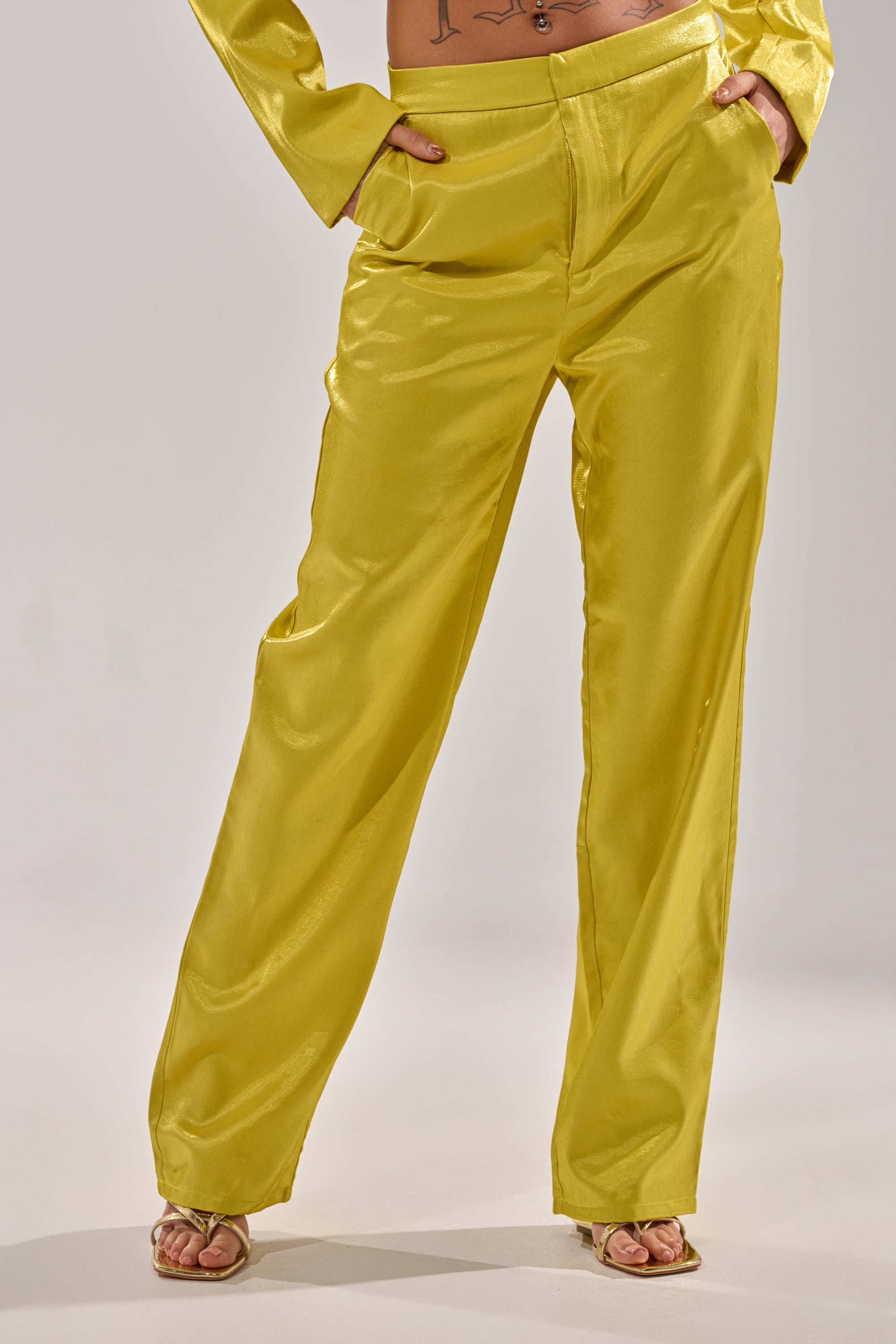 Model wears the CLEO TROUSER IN CHARTREUSE, featuring a high-rise waist and shiny finish, paired with a matching long-sleeve top. The look is styled with open-toe heels; image shows midsection and legs against a plain background. 1