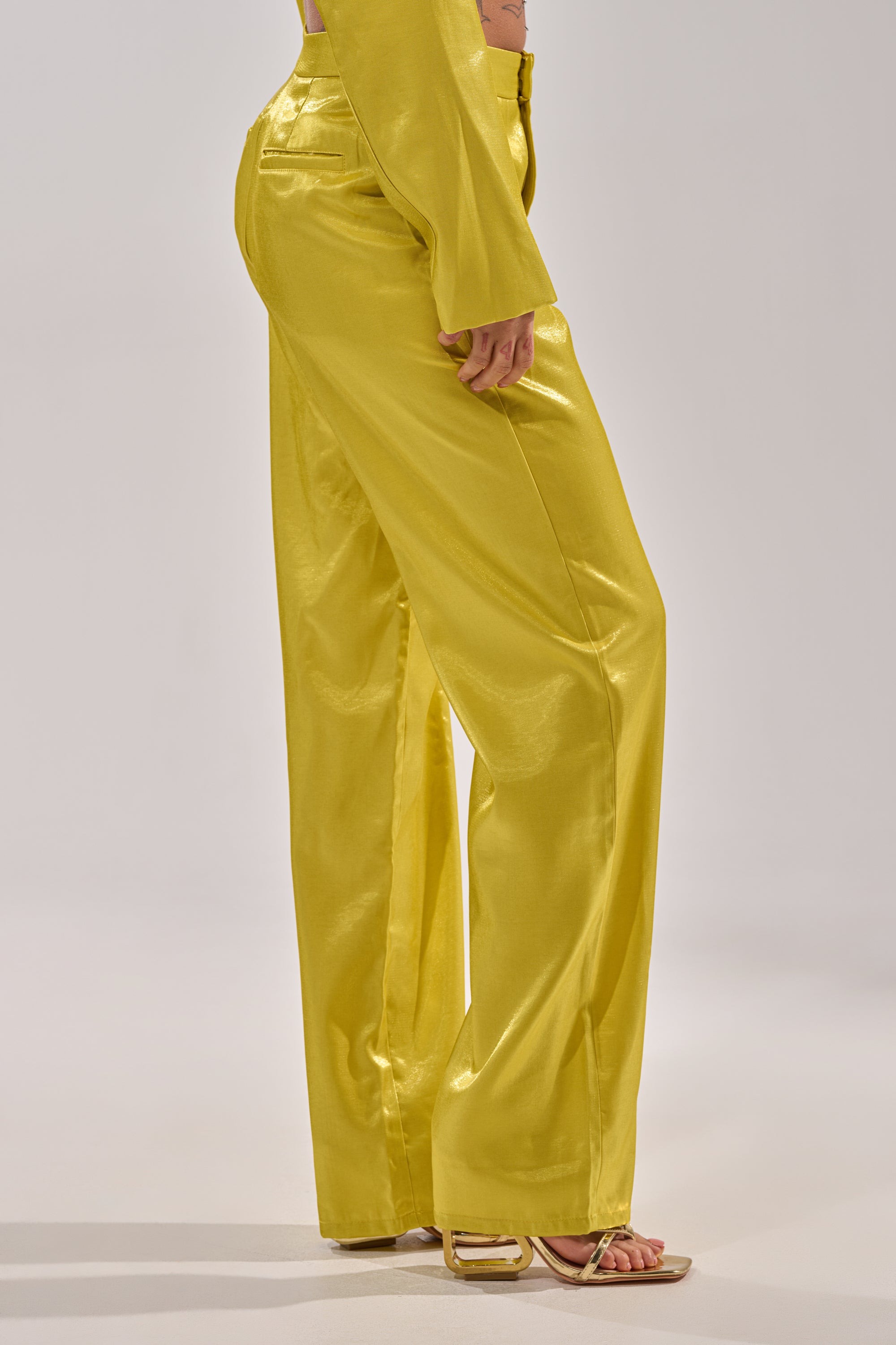 Model wears the CLEO TROUSER IN CHARTREUSE—shiny, high-rise, wide-leg pants with a matching long-sleeve top and open-toe heels. Side view showcases the fit and luxe fabric. 2