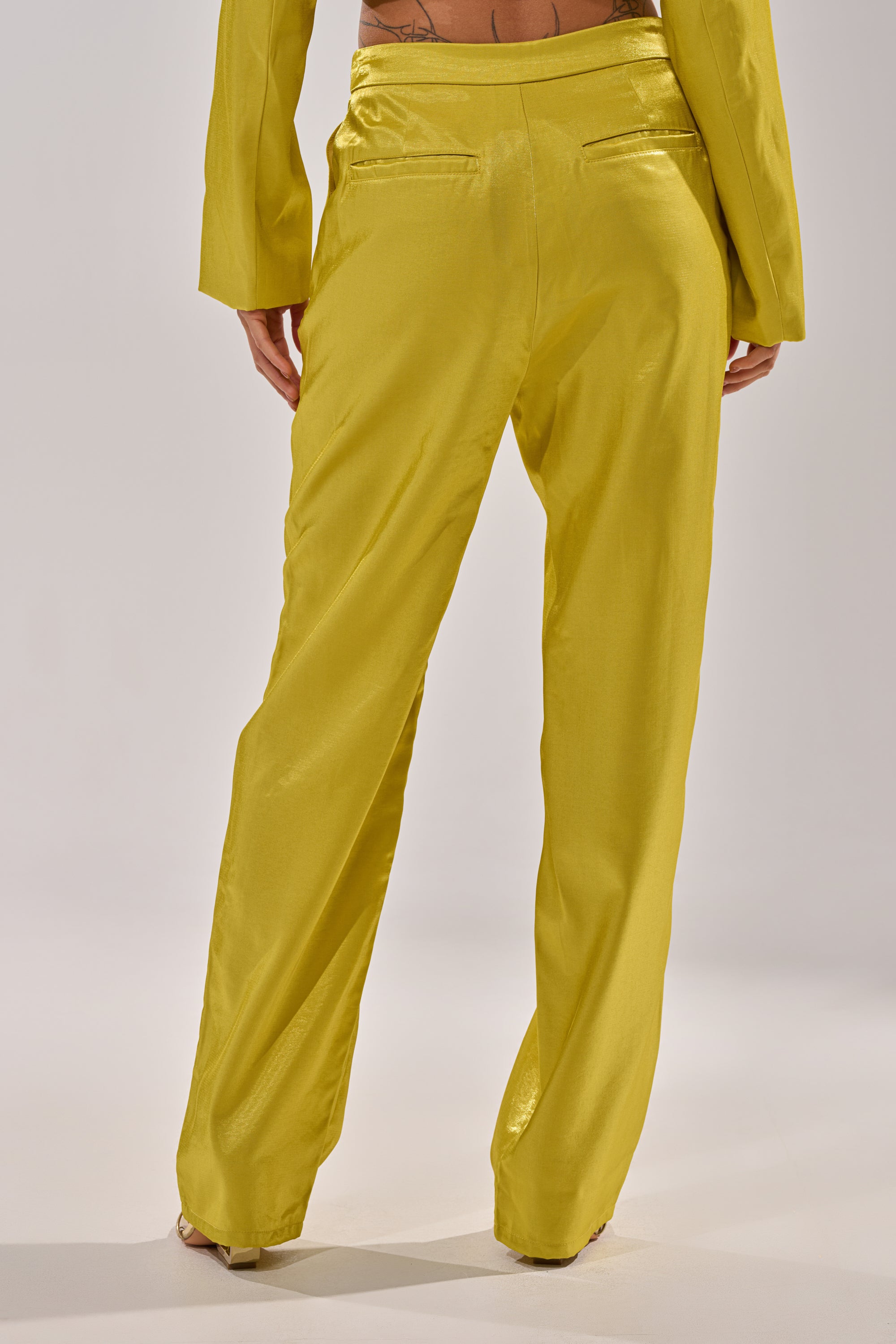 A person stands on a light floor, shown from the back with hands at their sides, wearing CLEO TROUSER IN CHARTREUSE with a matching yellow jacket; the straight-leg pants create an eye-catching look. 3