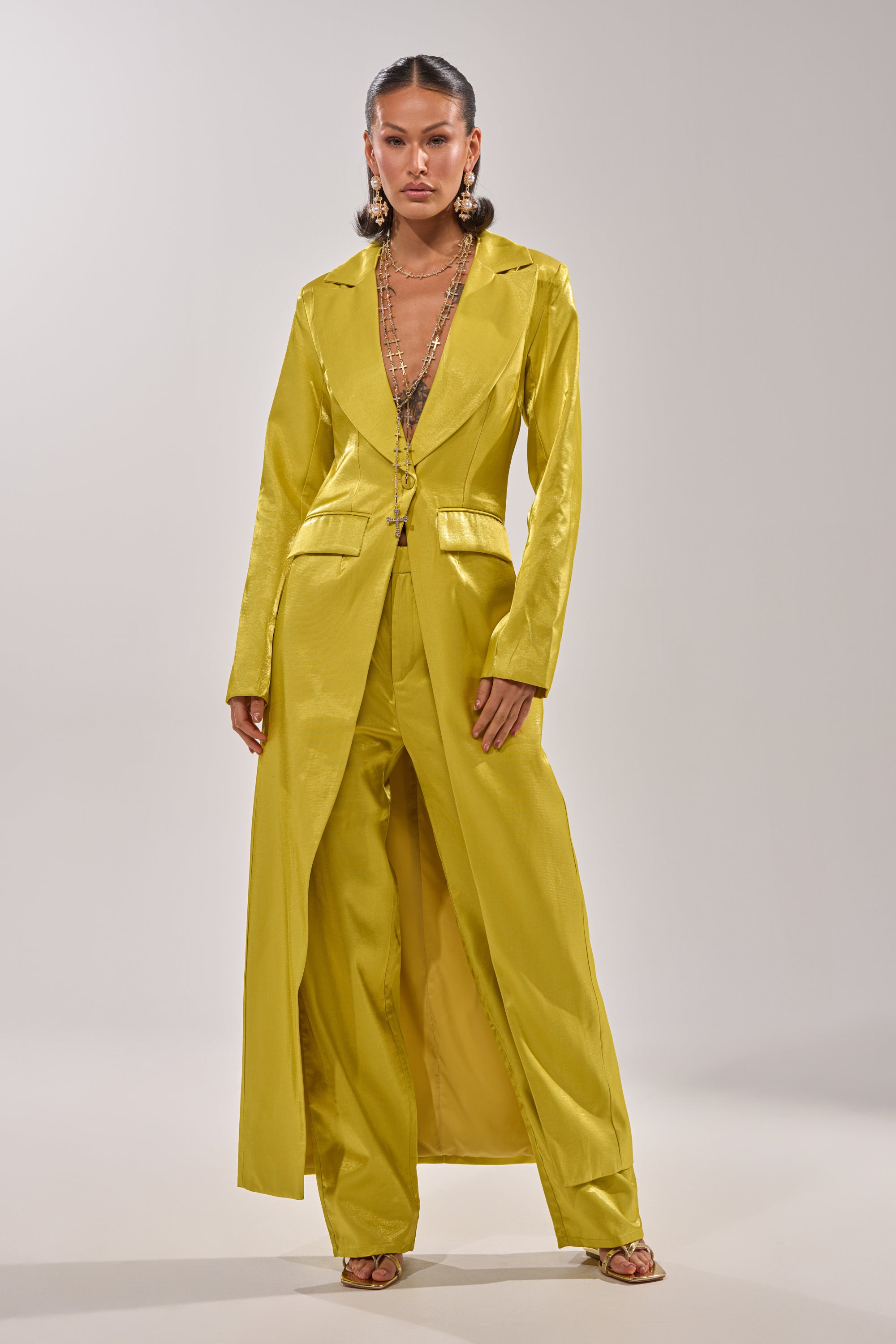 A woman stands confidently in CLEO TROUSER IN CHARTREUSE, a pair of high-rise, straight-leg pants, styled with a shiny yellow blazer, gold sandals, layered necklaces, and statement earrings against a light background. 5