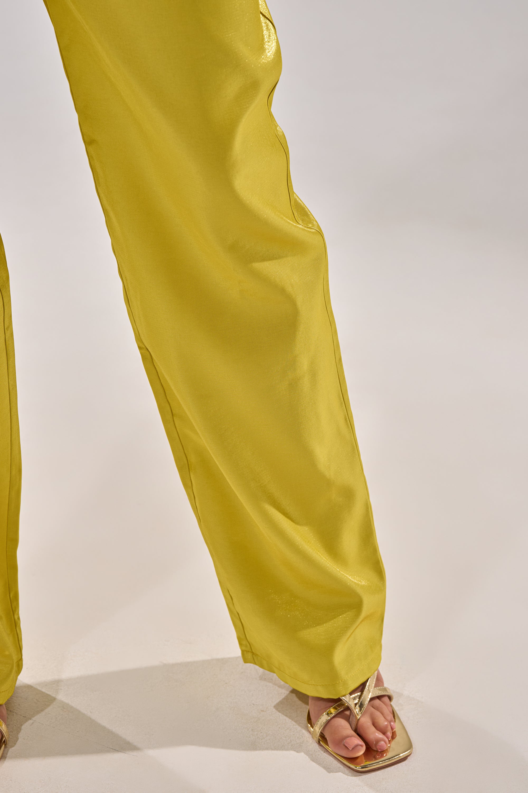 A person wears the CLEO TROUSER IN CHARTREUSE, featuring a high-rise waist, paired with gold open-toe sandals; only their lower legs and feet are shown on a light-colored surface. 7
