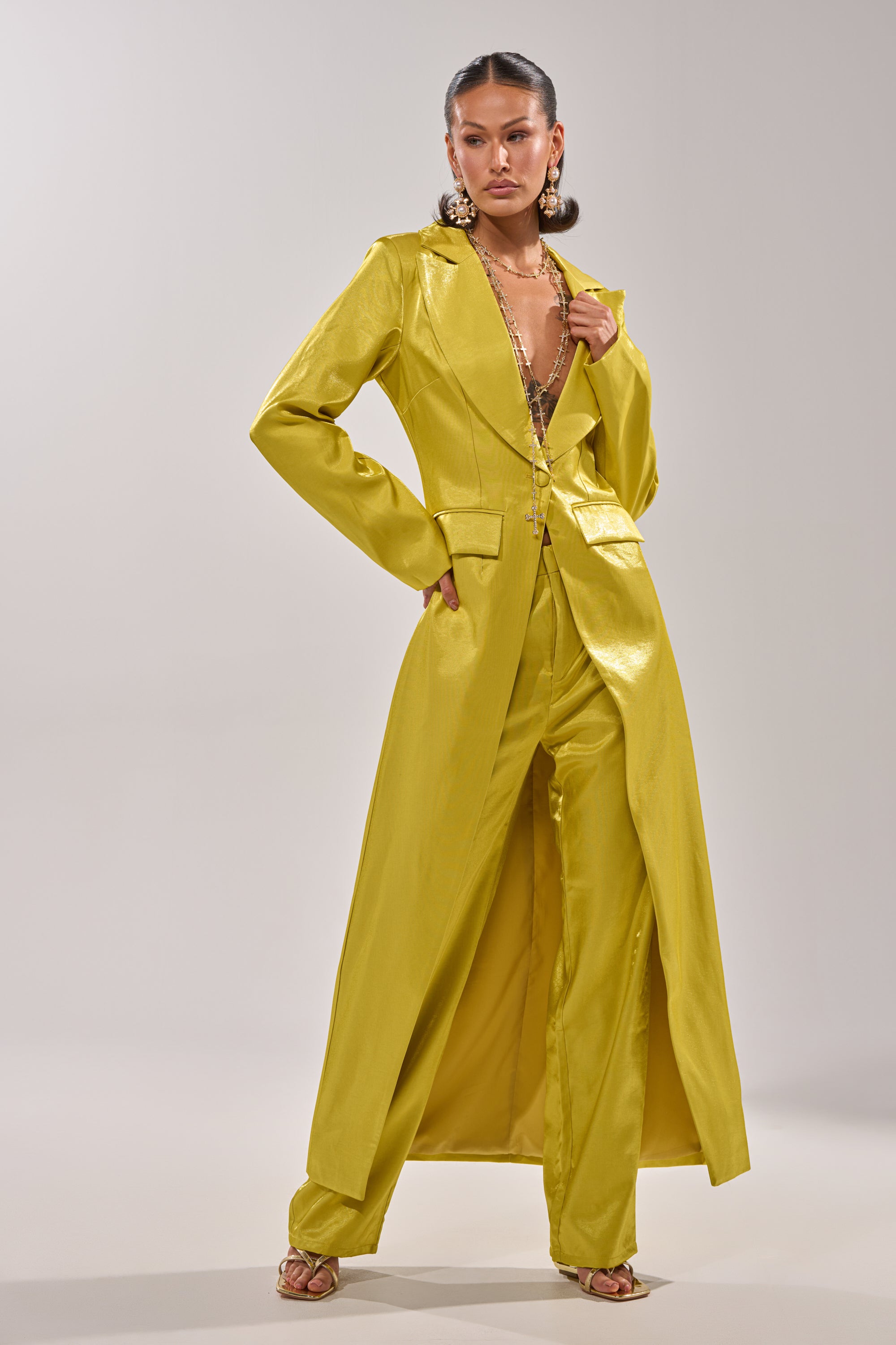 A person poses confidently in the CLEO TRENCH IN CHARTREUSE with matching wide-leg pants, styled with strappy heels, statement earrings, and layered necklaces against a neutral background. 1