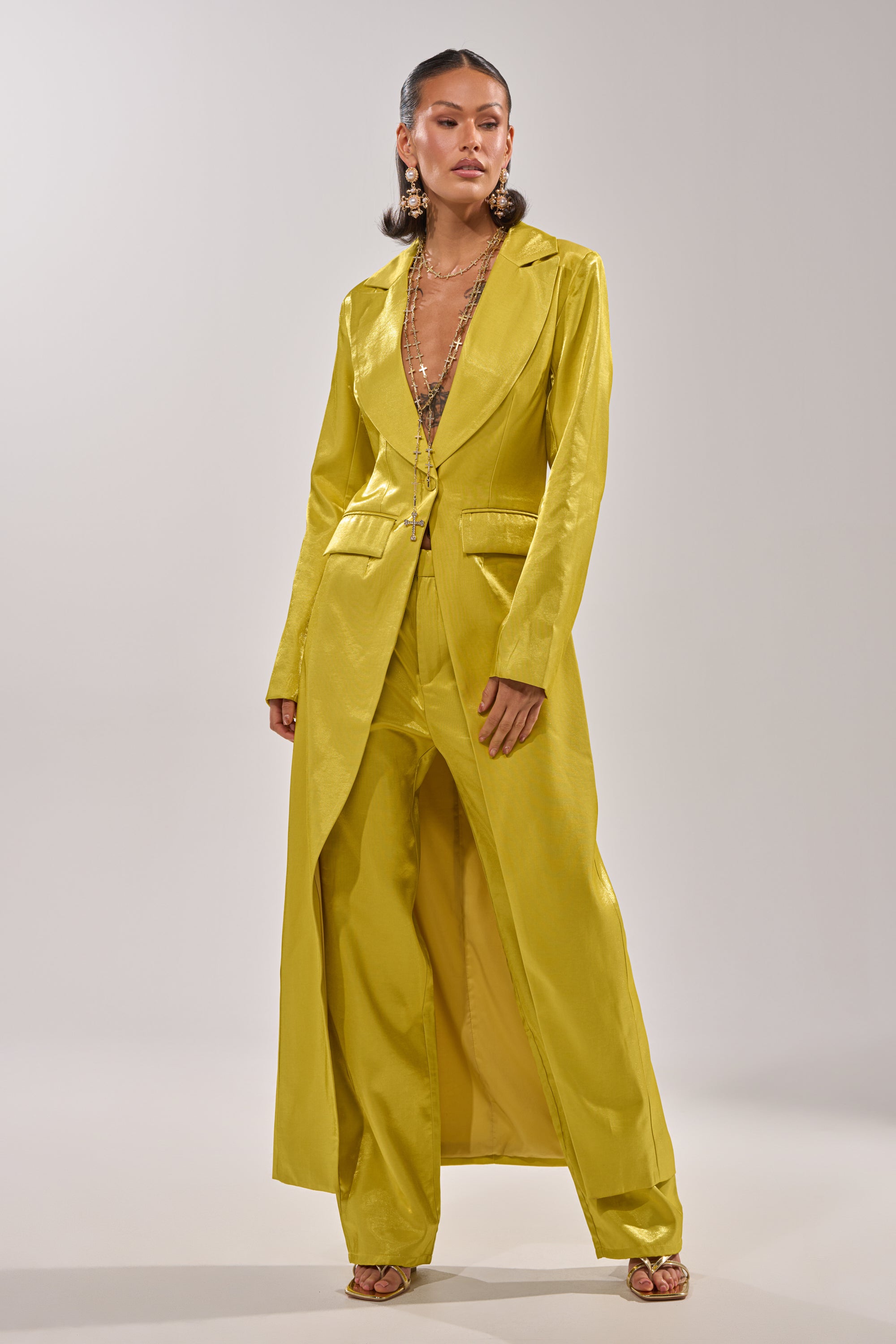 A woman models the CLEO TRENCH IN CHARTREUSE—featuring wide-legged pants and a long blazer—styled with gold earrings, layered necklaces, and slicked-back hair against a plain light background. 2