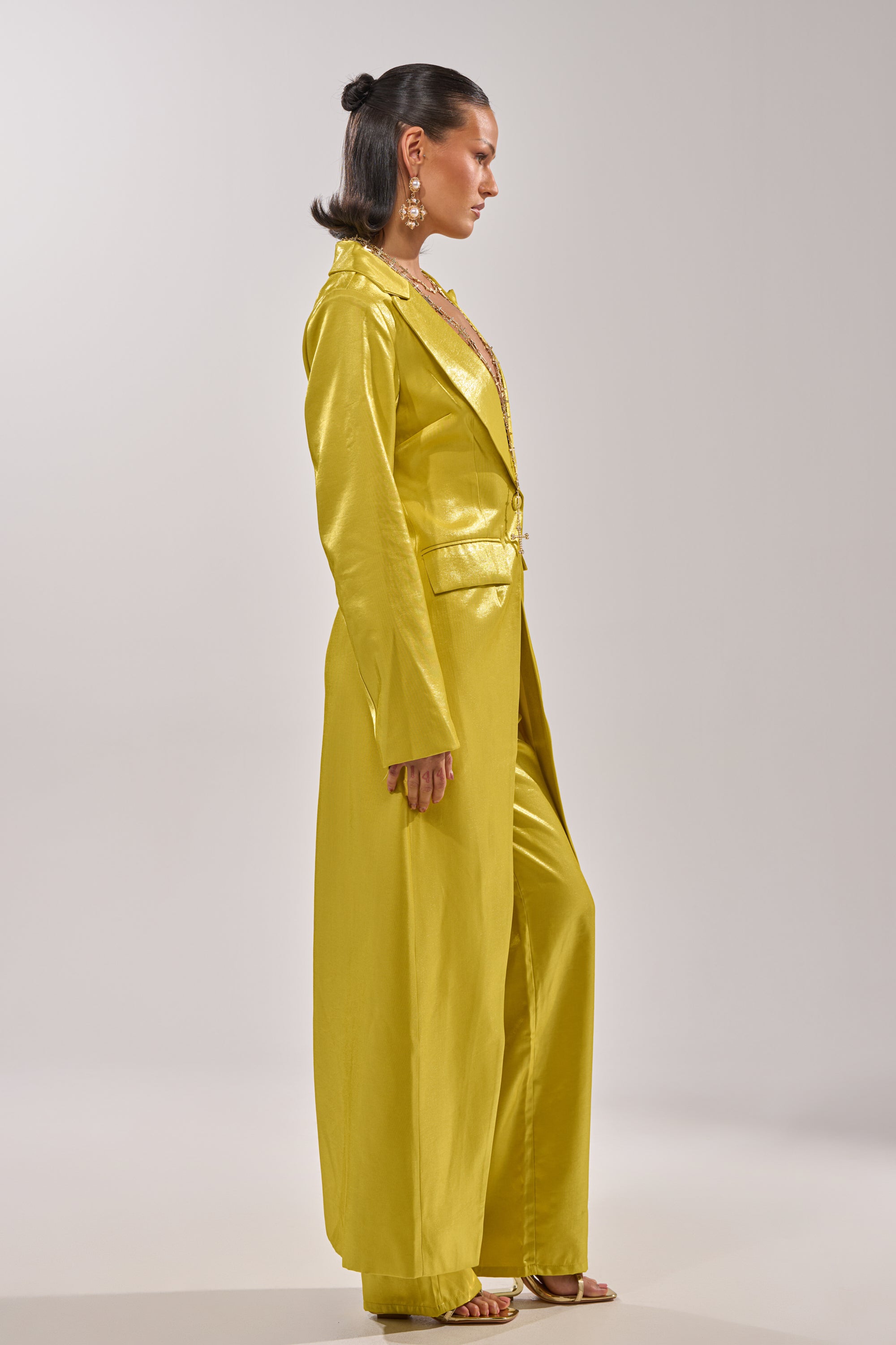 A woman stands sideways wearing the CLEO TRENCH IN CHARTREUSE with a structured silhouette, matching wide-leg pants, gold sandals, and statement earrings against a plain light background. 3