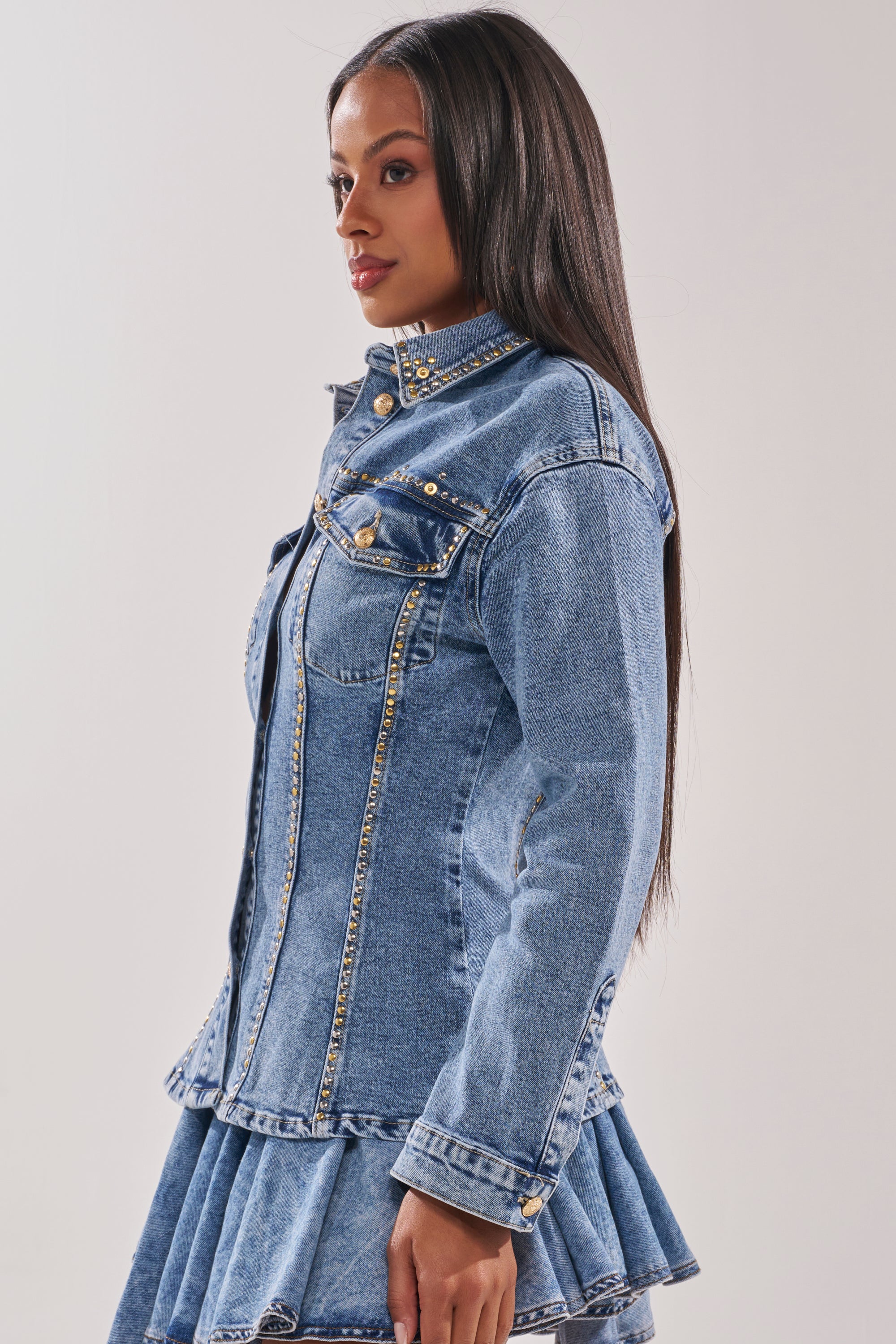 A woman with long straight hair wears the SHOULD'VE KNOWN BETTER STUDDED DENIM JACKET with gold-tone hardware and a matching pleated skirt, posing in profile against a plain light background. 2
