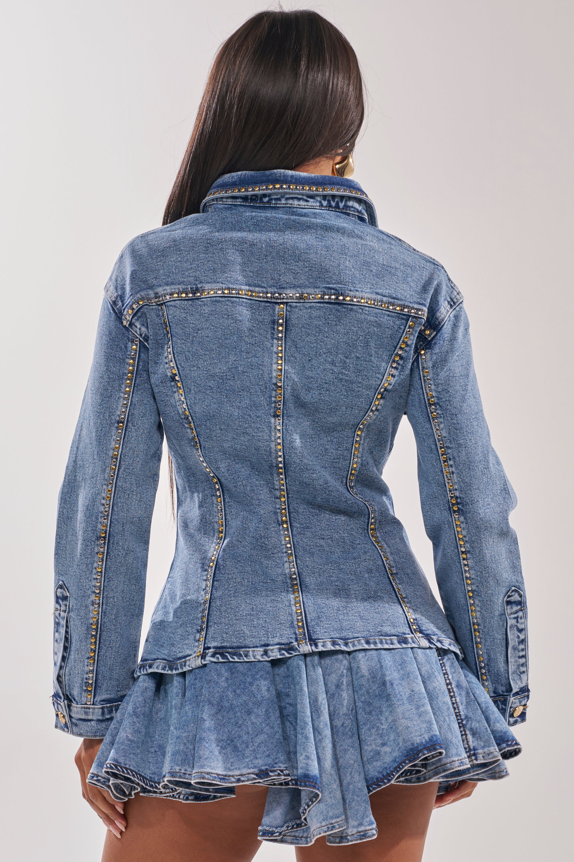 A woman with long dark hair is shown from the back, wearing the SHOULD'VE KNOWN BETTER STUDDED DENIM JACKET with gold-tone hardware and a short, pleated denim skirt. The background is plain light gray. 3