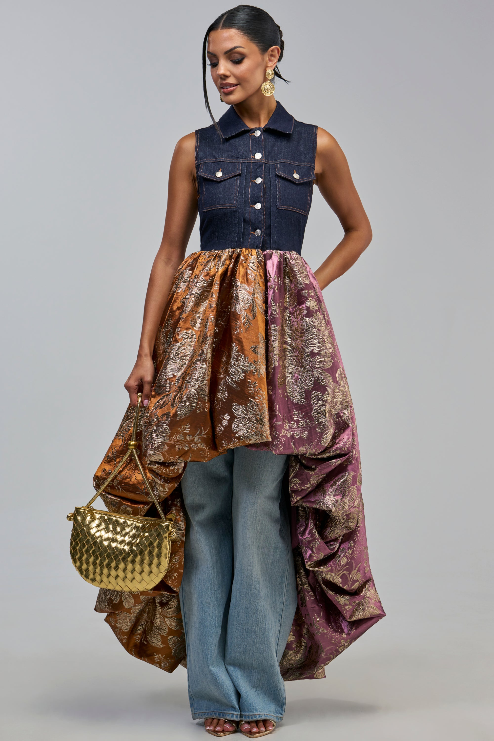 A model wearing the ON THE HIGH LOW DENIM VEST IN DARK BLUE DENIM paired with a bronze and purple floral bubble skirt over light-wash jeans, carrying a gold woven handbag.