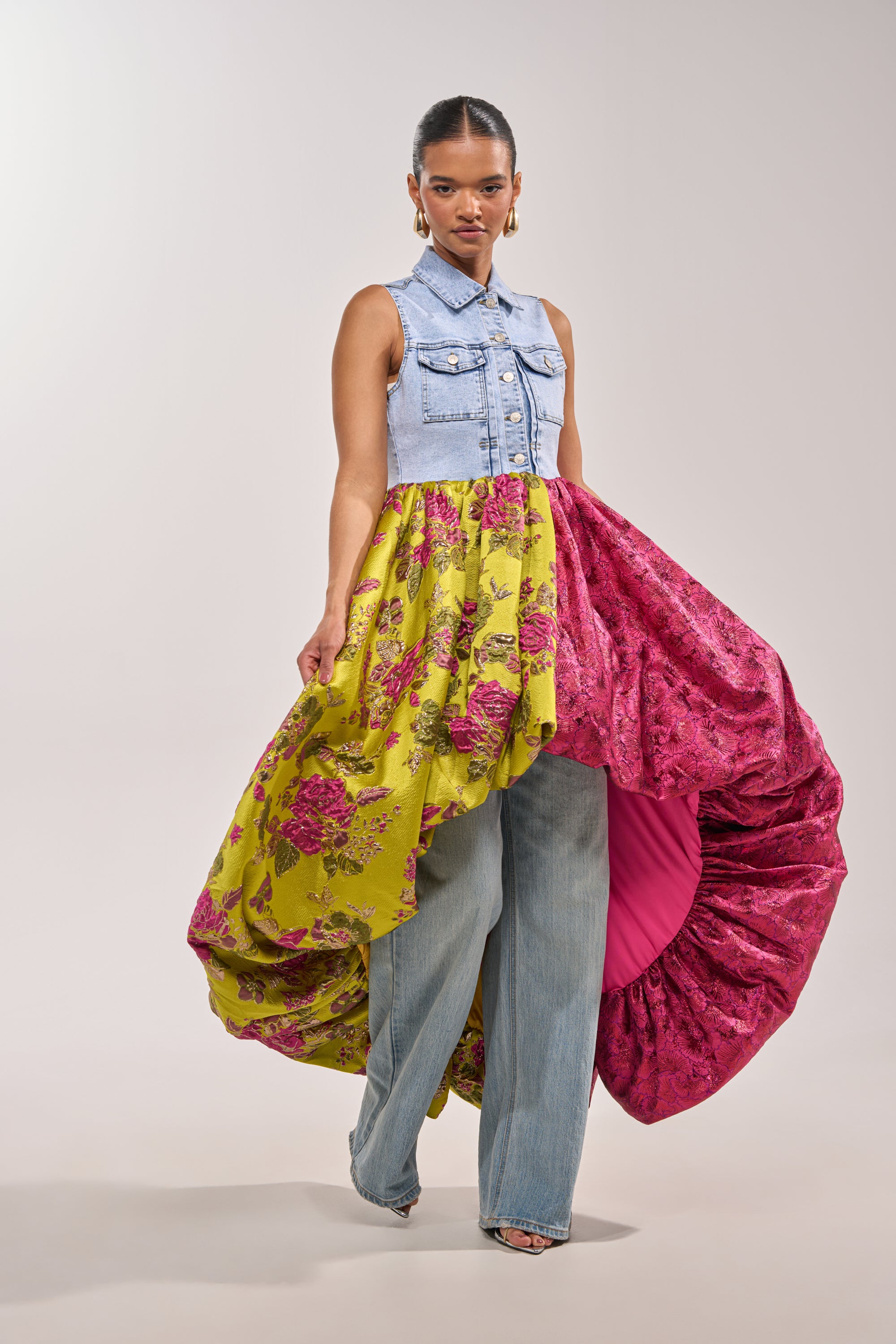 A woman models the ON THE HIGH LOW DENIM VEST, styled with a yellow floral brocade, pink ruffled skirt over blue jeans, large hoop earrings, and open-toe shoes—perfect for bold statement occasion wear against a plain background. 0
