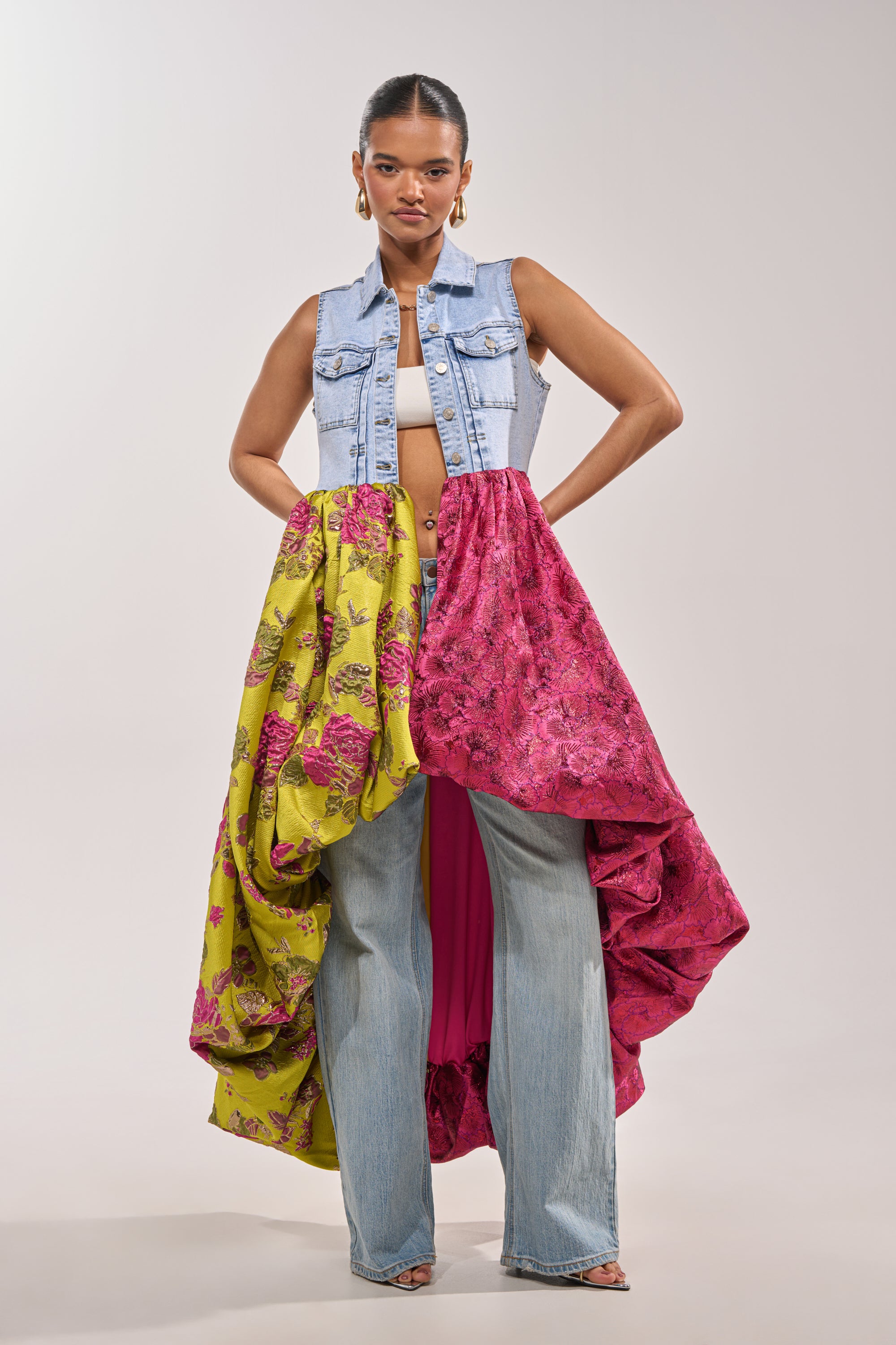A woman stands confidently in the ON THE HIGH LOW DENIM VEST, light blue jeans, a white crop top, and a vibrant yellow-and-pink floral skirt—a bold look ideal for festivals or standout occasions. 1