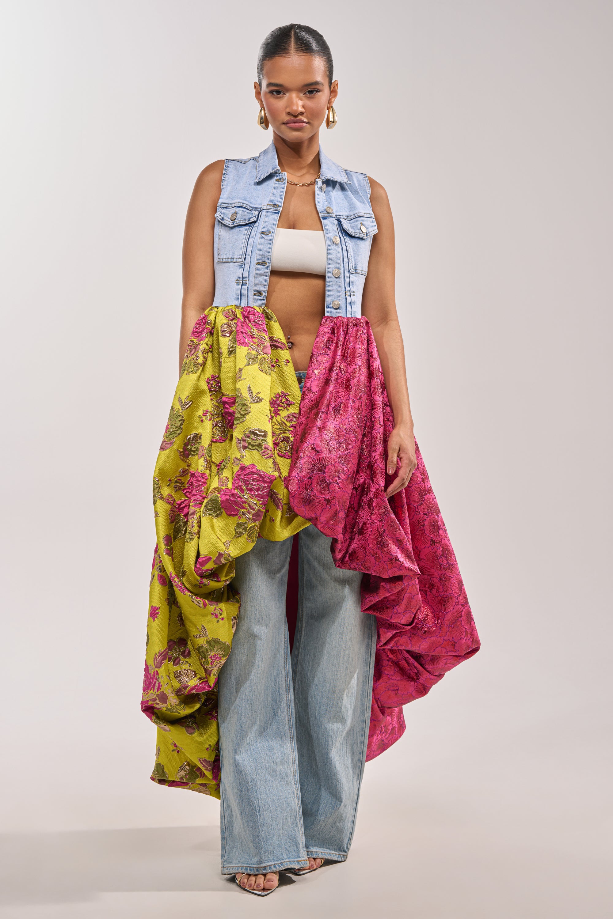 Wearing the ON THE HIGH LOW DENIM VEST over a white crop top, wide-leg jeans, and a bold skirt with yellow and magenta floral draping, she exudes festival-ready style—finished with hoop earrings and heels. 2
