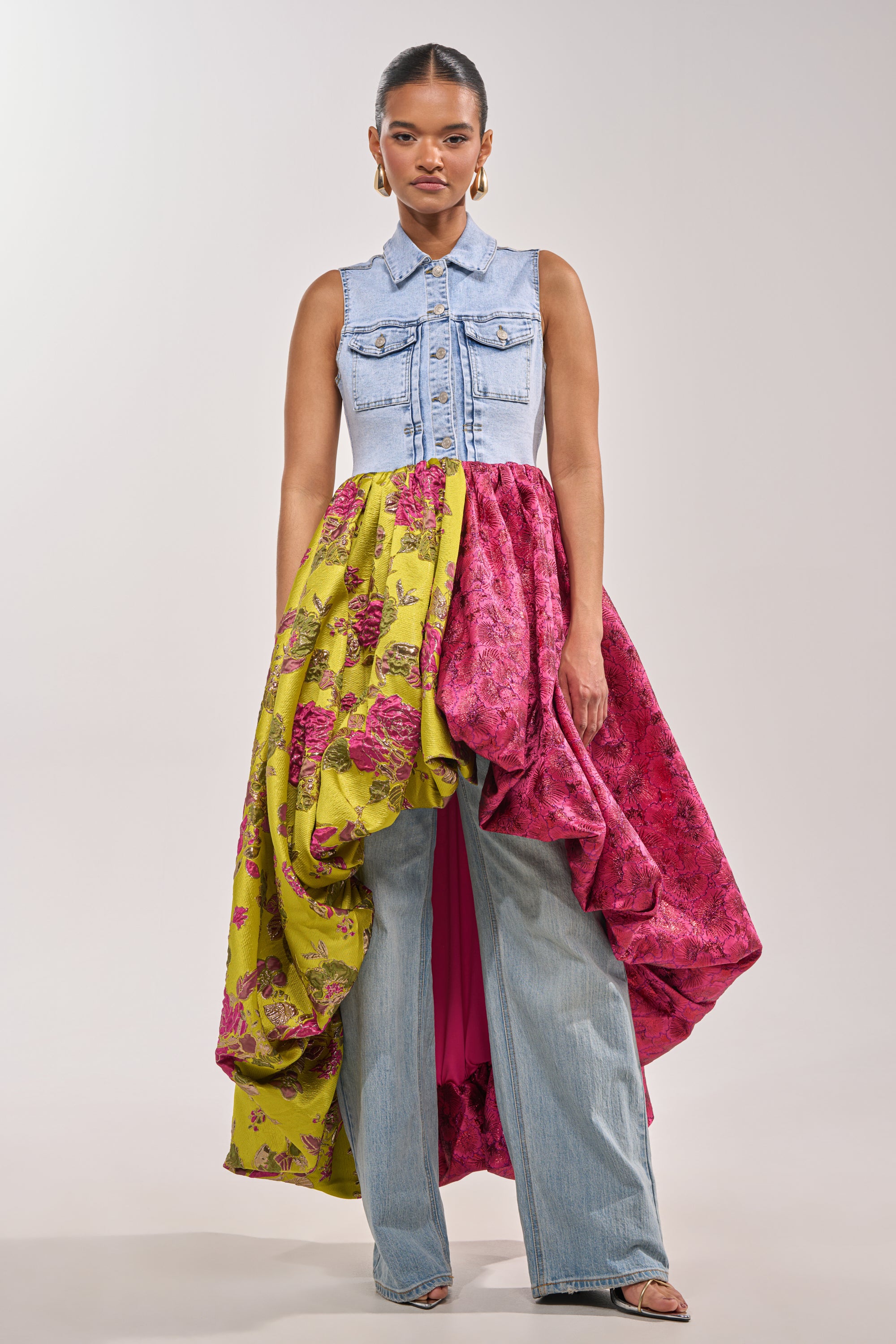A woman channels bold festival style in the ON THE HIGH LOW DENIM VEST and a yellow and pink floral skirt over flared blue jeans, standing against a plain light background. 3