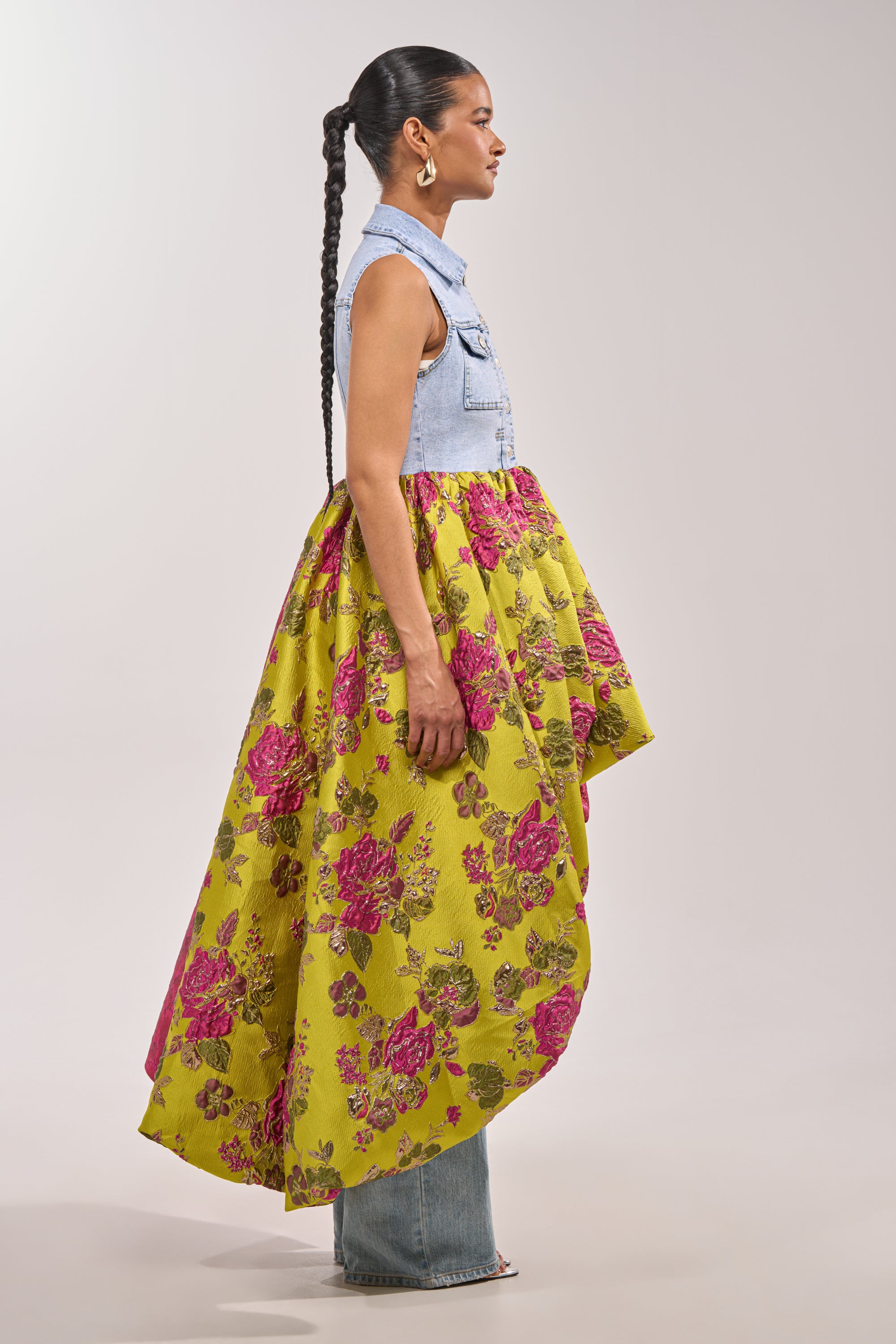 A woman with a long braid models the ON THE HIGH LOW DENIM VEST, paired with a yellow floral high-low skirt and blue jeans. She accessorizes with hoop earrings and stands in profile against a light background for fresh festival style. 4