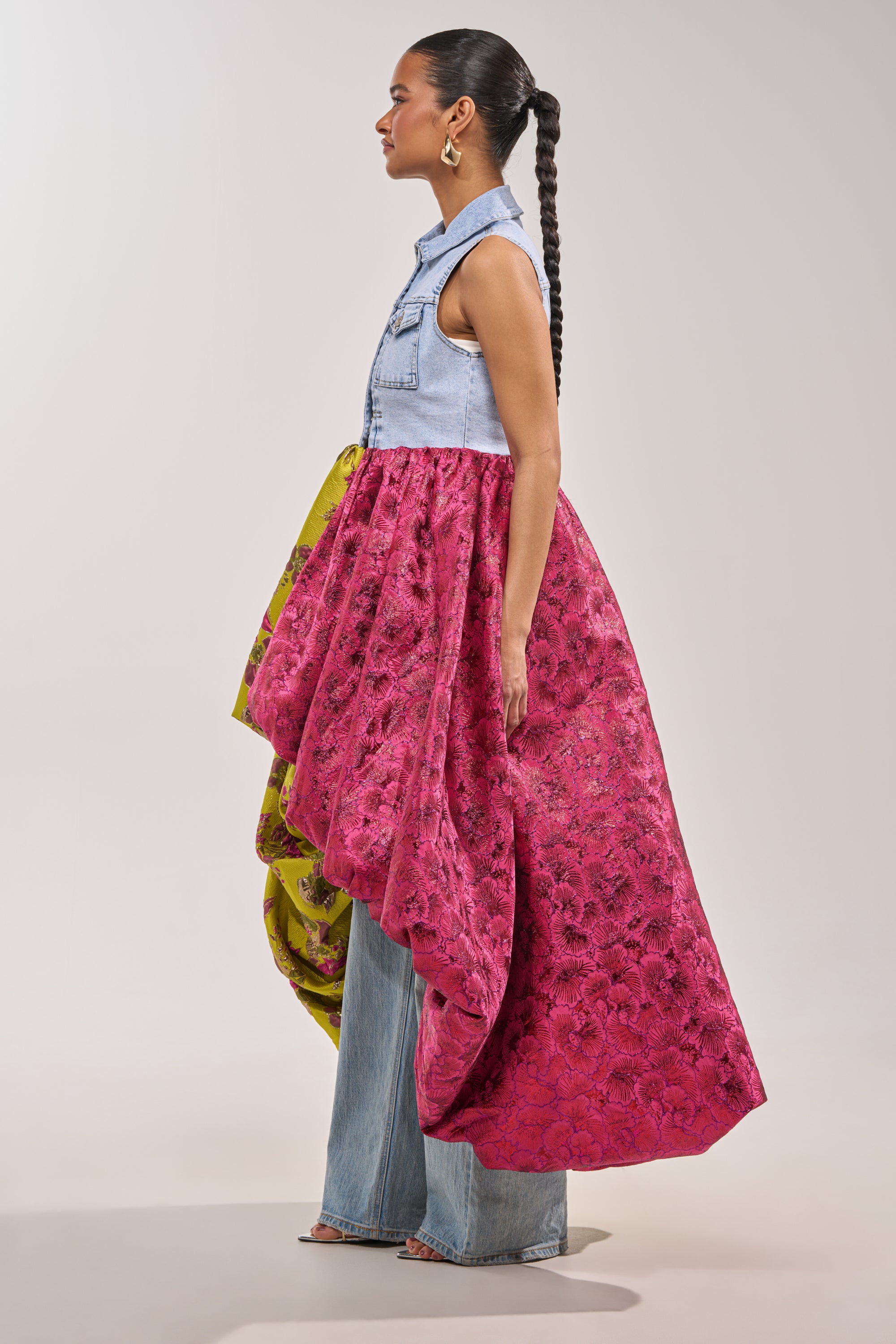 A woman stands in profile wearing the ON THE HIGH LOW DENIM VEST with an attached pink and yellow floral brocade skirt, paired with wide-leg denim pants and large gold hoop earrings for bold festival flair. 5