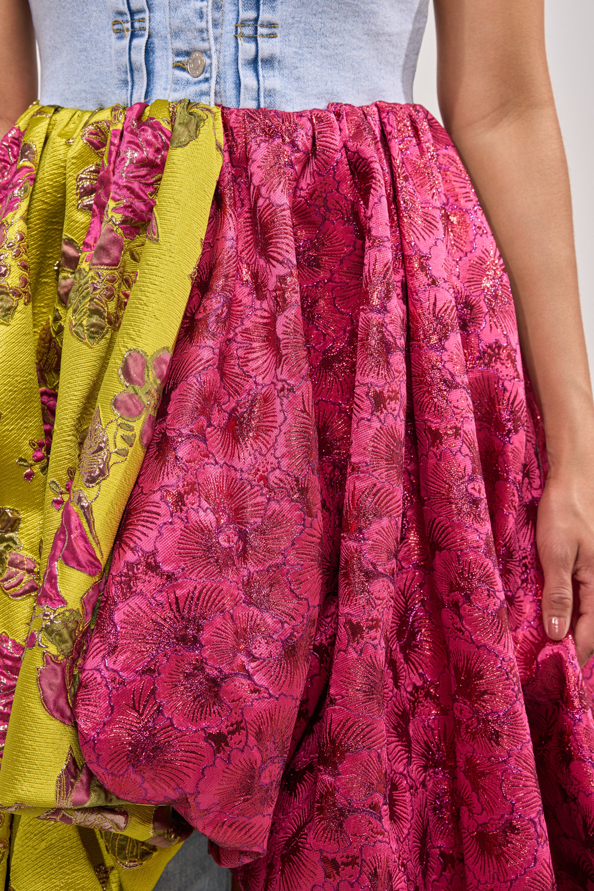 A close-up of a person wearing the ON THE HIGH LOW DENIM VEST, featuring a denim bodice and a voluminous yellow-green and pink floral brocade skirt, with their arm and hand partially visible. 7