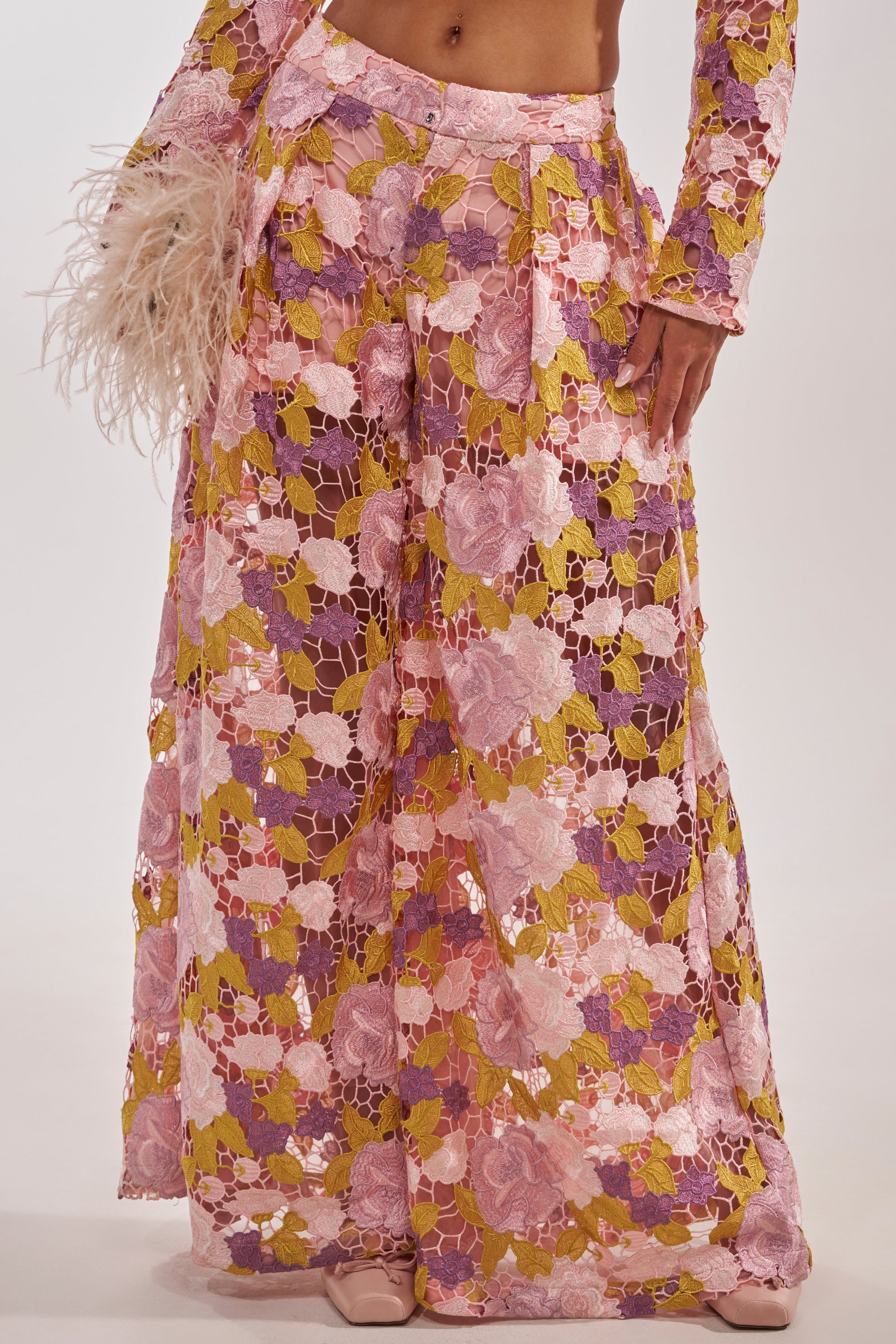 Wearing the LILA LACE PANT—embroidered floral trousers crafted from pink, purple, and yellow sheer lace—a hand holds a pink feather detail. 3