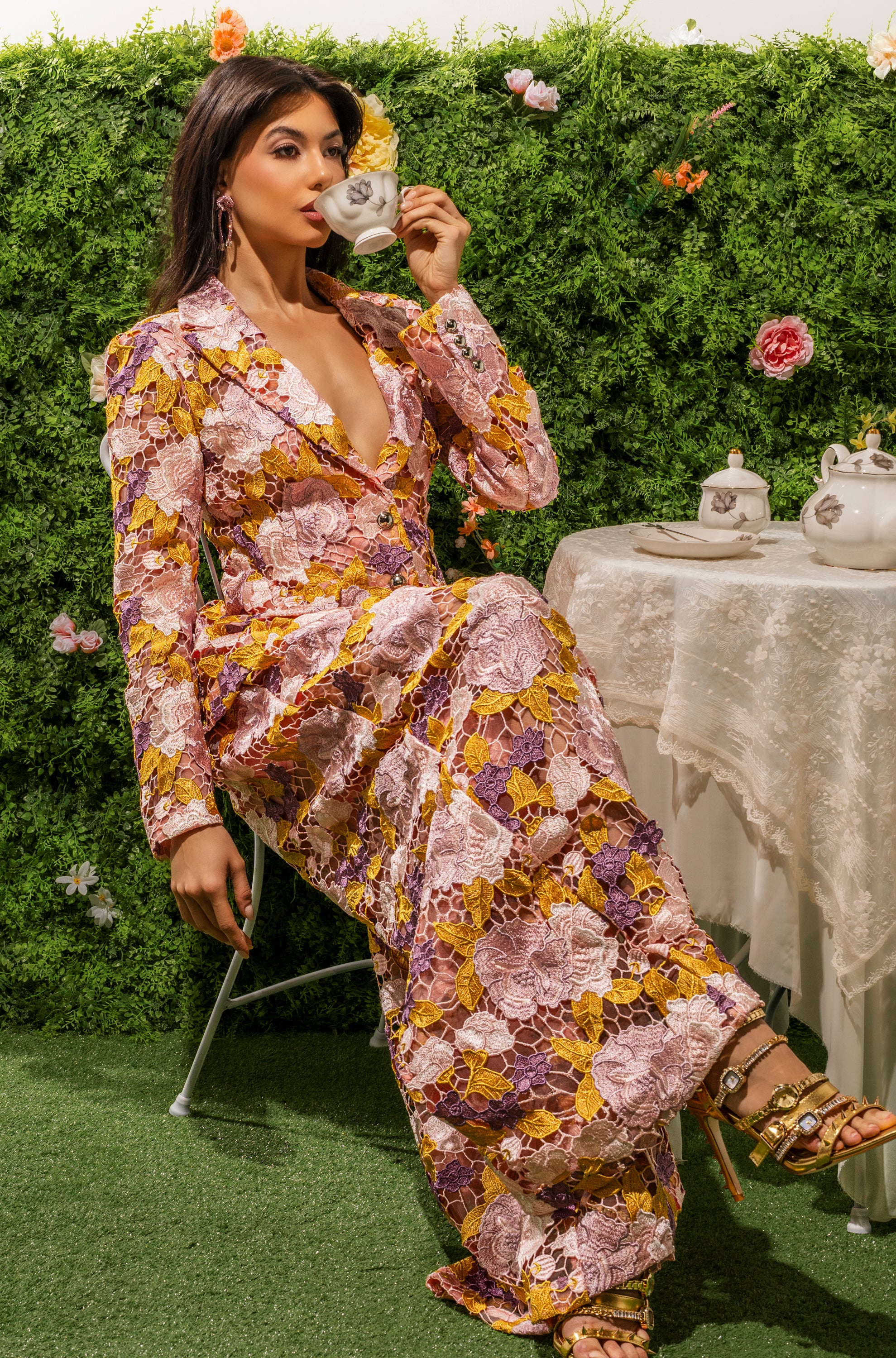 Wearing the LILA LACE PANT, a woman sits outdoors on a chair, elegantly sipping tea. A lush hedge and colorful flowers form her backdrop, while a lace-adorned table with an inviting tea set adds charm to the scene. 1