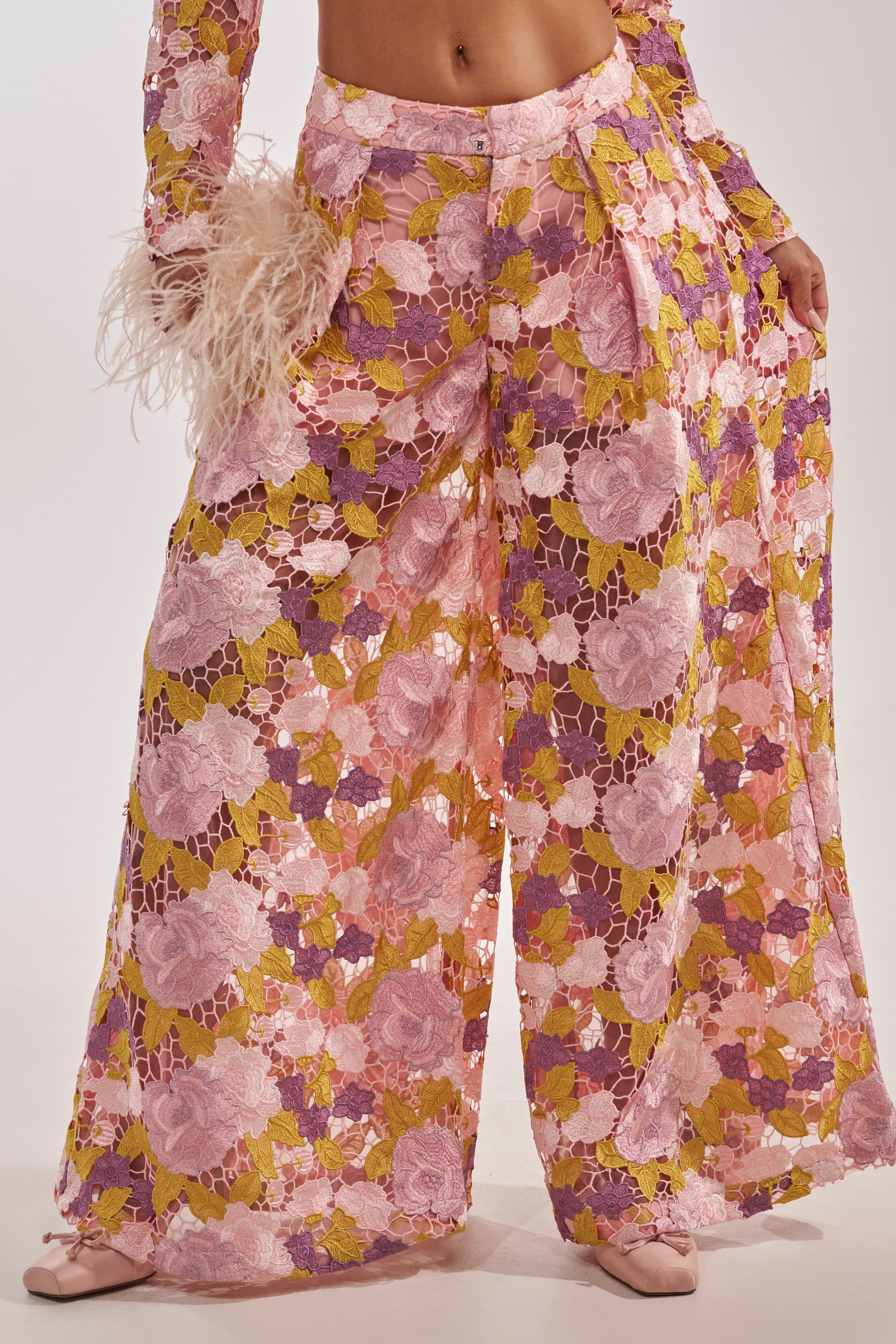 A person wears the LILA LACE PANT, a vintage-luxe wide-leg trouser featuring a pink, purple, and yellow floral lace pattern. Styled with a matching sleeve trimmed in pink feathers, they pose against a plain backdrop. 4