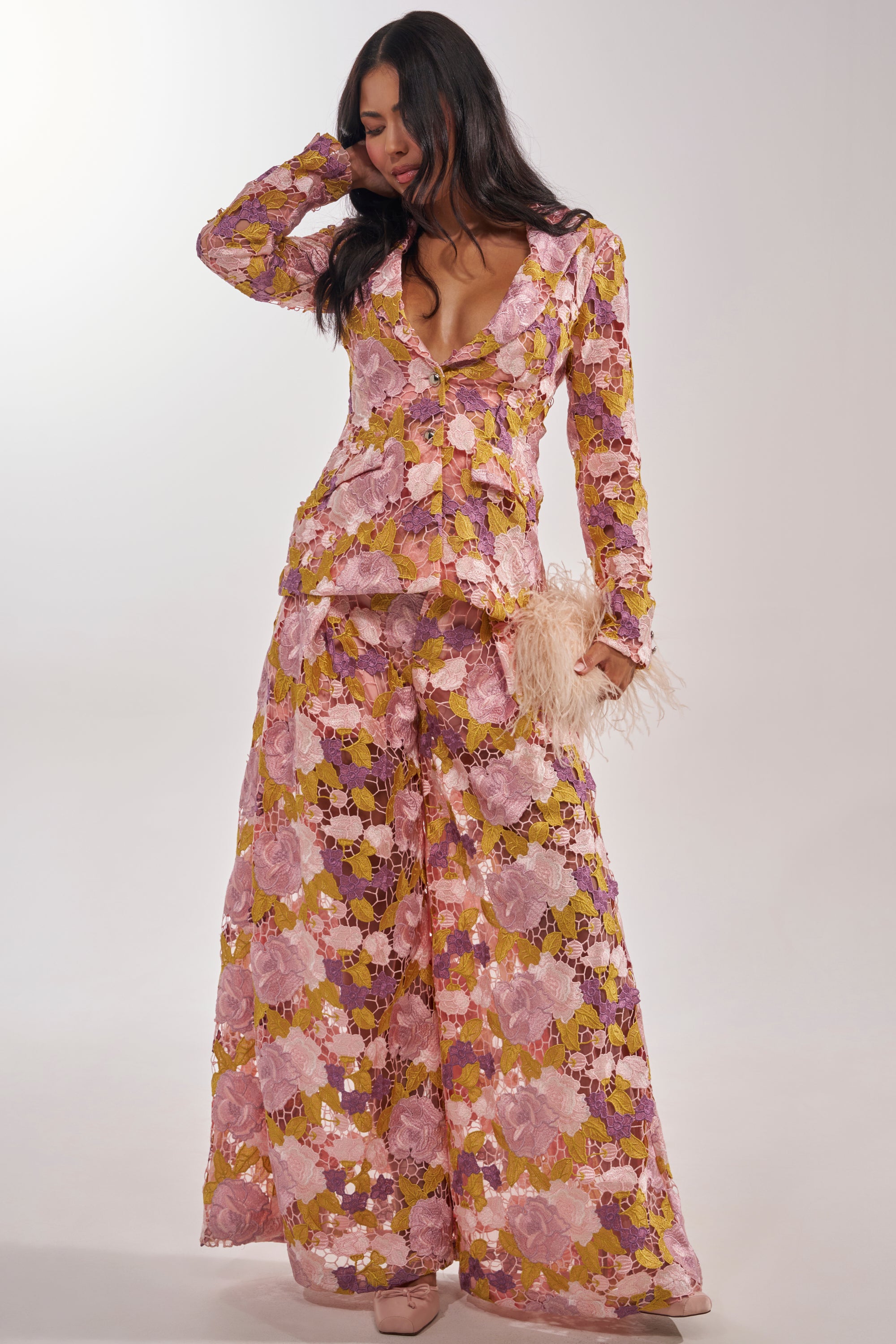 A woman showcases the LILA LACE PANT, a vintage-luxe floral lace pantsuit in pink, yellow, and purple. She accessorizes with a feathered clutch and nude heels, wearing wide-leg trousers against a plain white backdrop. 7